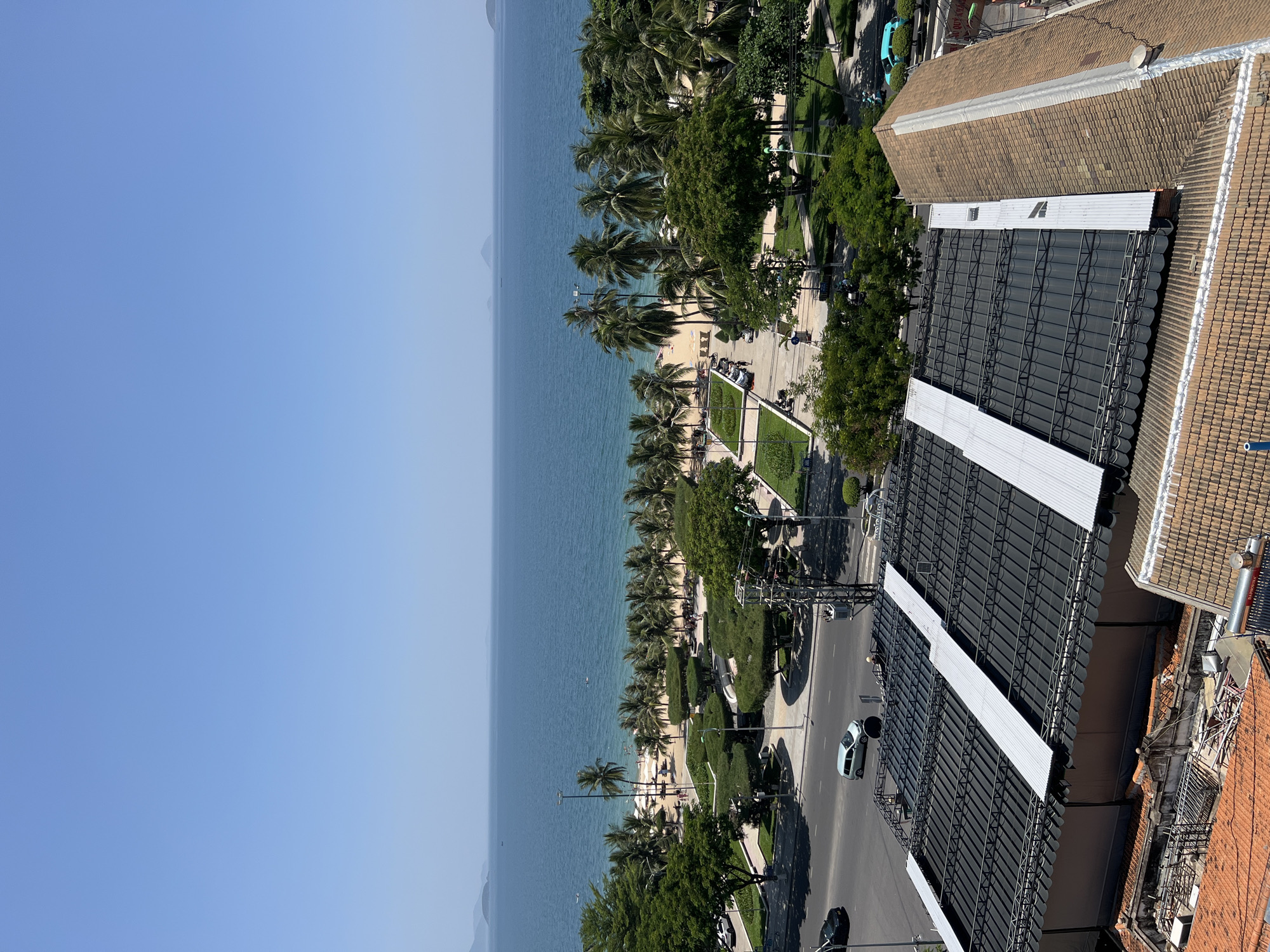 The image depicts a daytime aerial view of a coastal city, likely Nha Trang, Vietnam, overlooking a beachfront and the ocean. In the foreground, a building with a dark, corrugated metal roof featuring several lighter-colored metal strips is prominent. Adjacent to this is another building with a brown, tiled roof. Below these structures, a street is visible with a silver car driving and a black car parked. Beyond the street lies a landscaped area with manicured trees and walkways. Further back, a sandy beach is lined with numerous palm trees and scattered beachgoers. The ocean stretches to the horizon, with a clear blue sky overhead and distant islands visible. The overall scene is characteristic of a tropical urban environment with a focus on the coastline.