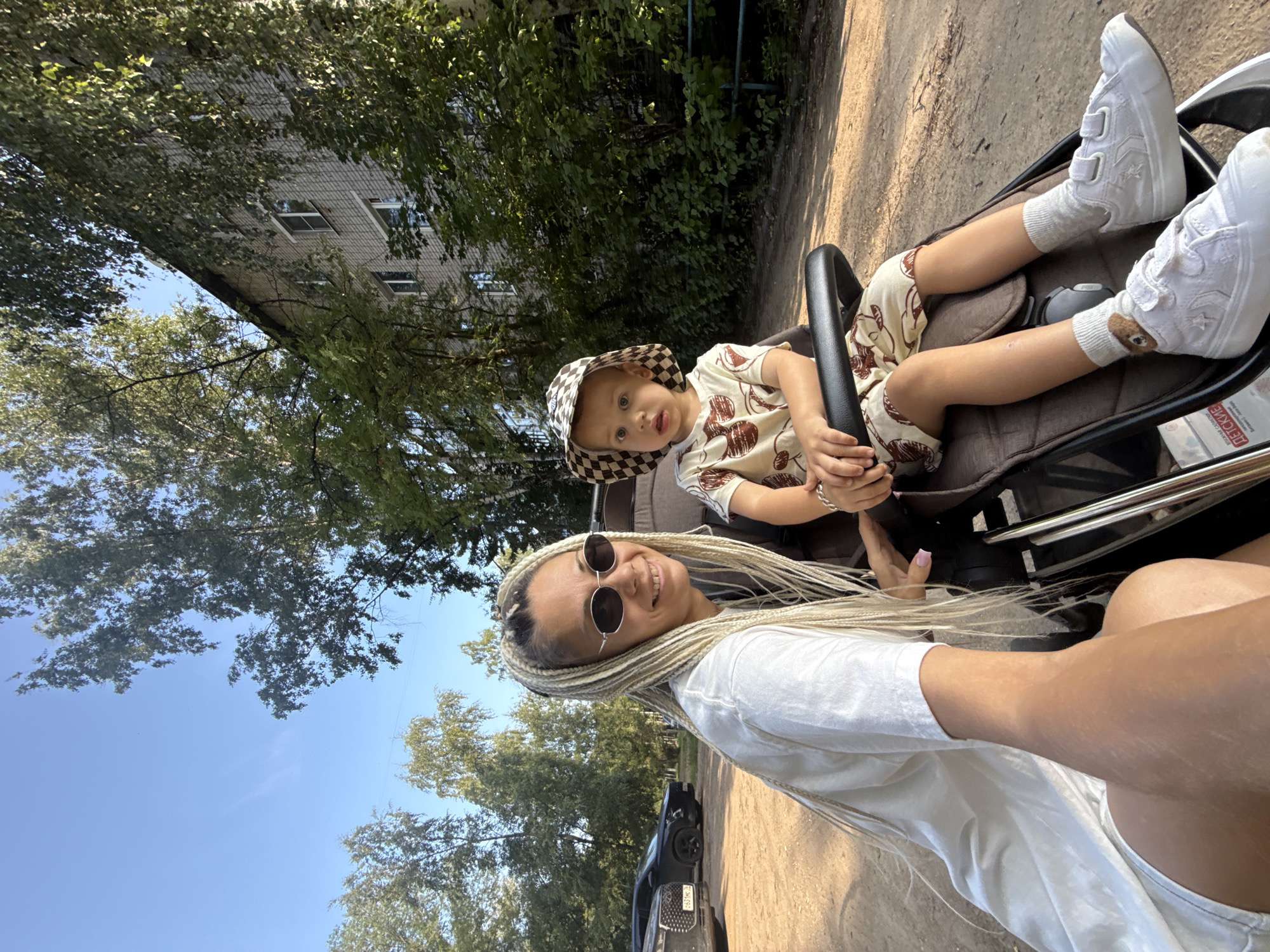 A medium-shot, outdoor, candid photograph shows a woman and a child in a stroller on a sunny day. The woman, who is to the left of the frame, has long blonde braided hair and wears sunglasses and a white t-shirt. She is smiling at the camera and her left arm is extended towards the camera, as if taking a selfie. The child, who is in the stroller to the right, is wearing a patterned hat and a matching outfit. The child is looking away from the camera with a surprised expression.
The background of the image shows a tree-lined street with a multi-story apartment building in the distance. The ground is covered with gravel, suggesting a rural or suburban setting. A black car is parked to the left of the frame. The scene appears to be in the city of Rusanovo, Russia. The lighting and shadows indicate it is late afternoon.