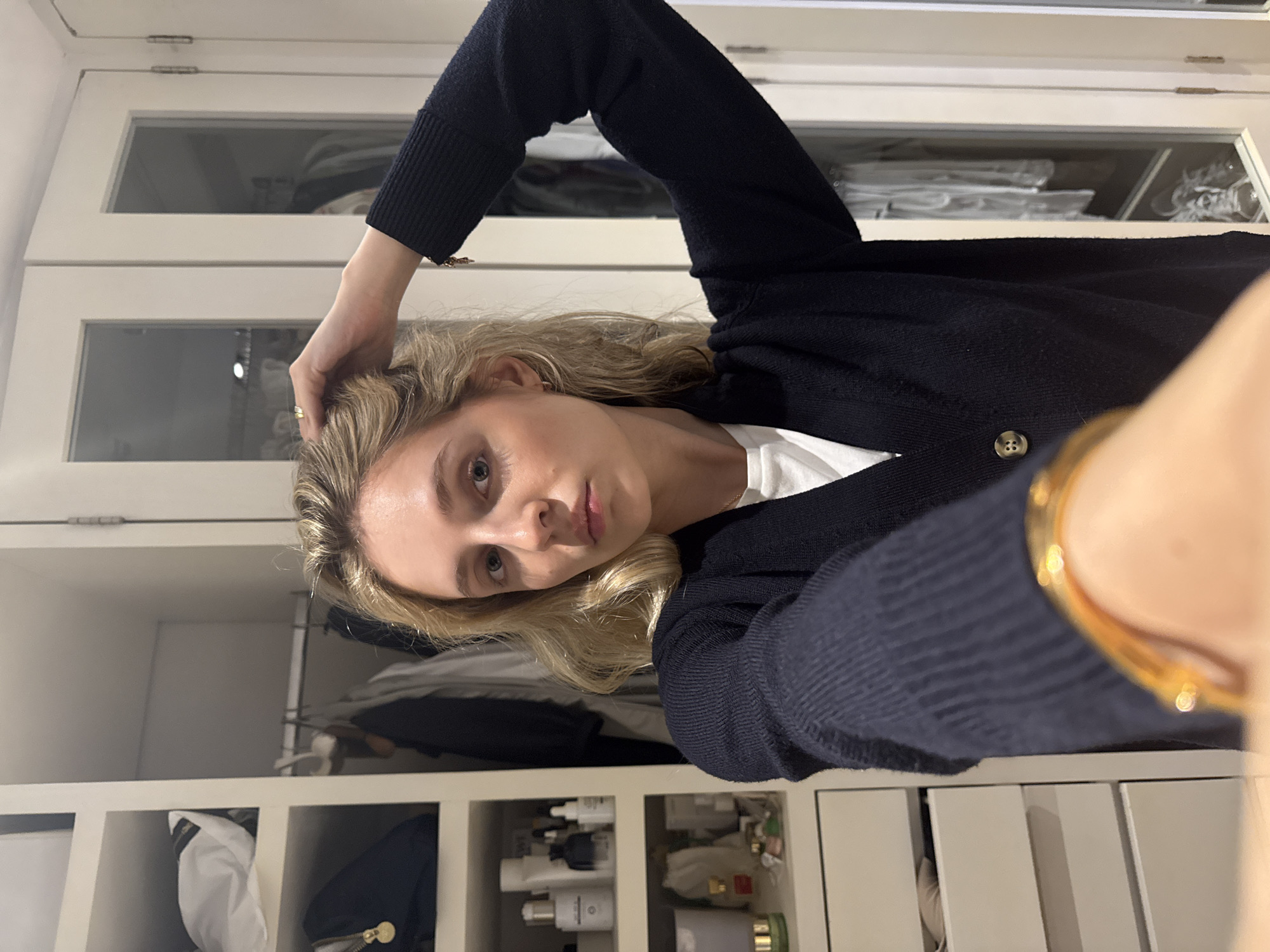 A young woman with blonde hair, wearing a dark blue cardigan over a white t-shirt, is standing in front of a closet. Her right hand is raised, touching her hair, and her left arm is bent, with her hand resting on her hip. She is looking directly at the camera. The closet has white doors and shelves filled with clothes and various products. The scene appears to be indoors, possibly a bedroom or dressing room. There is no visible indication of the location being Mexico City.