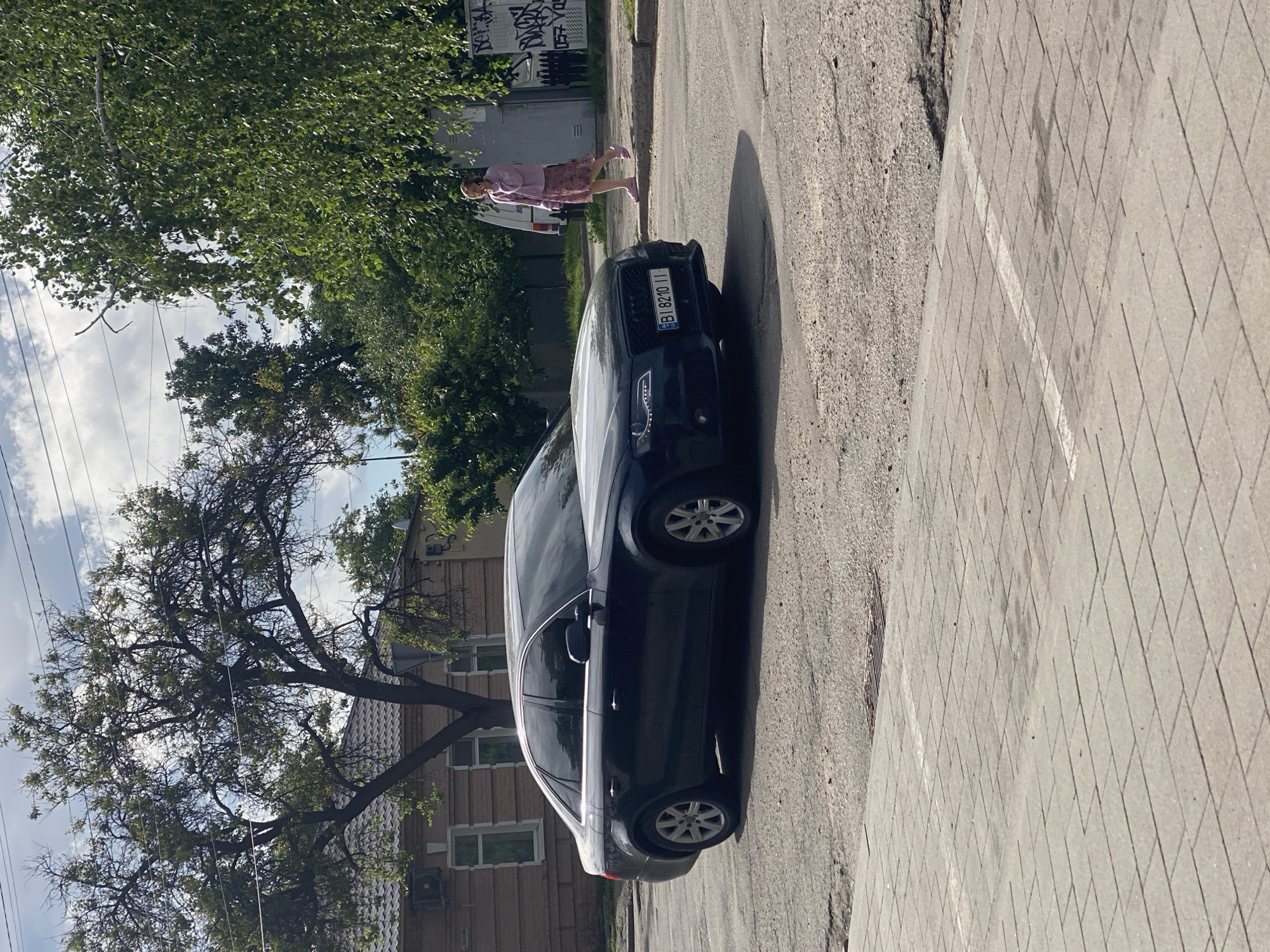 The image shows a black Audi parked on a street in Poltava, Ukraine. A person in pink clothes stands on the sidewalk near the car. The car's license plate is visible, reading "BI 8240 II". Trees line the street, and a building can be seen in the background. The street surface is asphalt, and the sidewalk is paved with light-colored tiles. Overhead power lines are visible in the sky.
