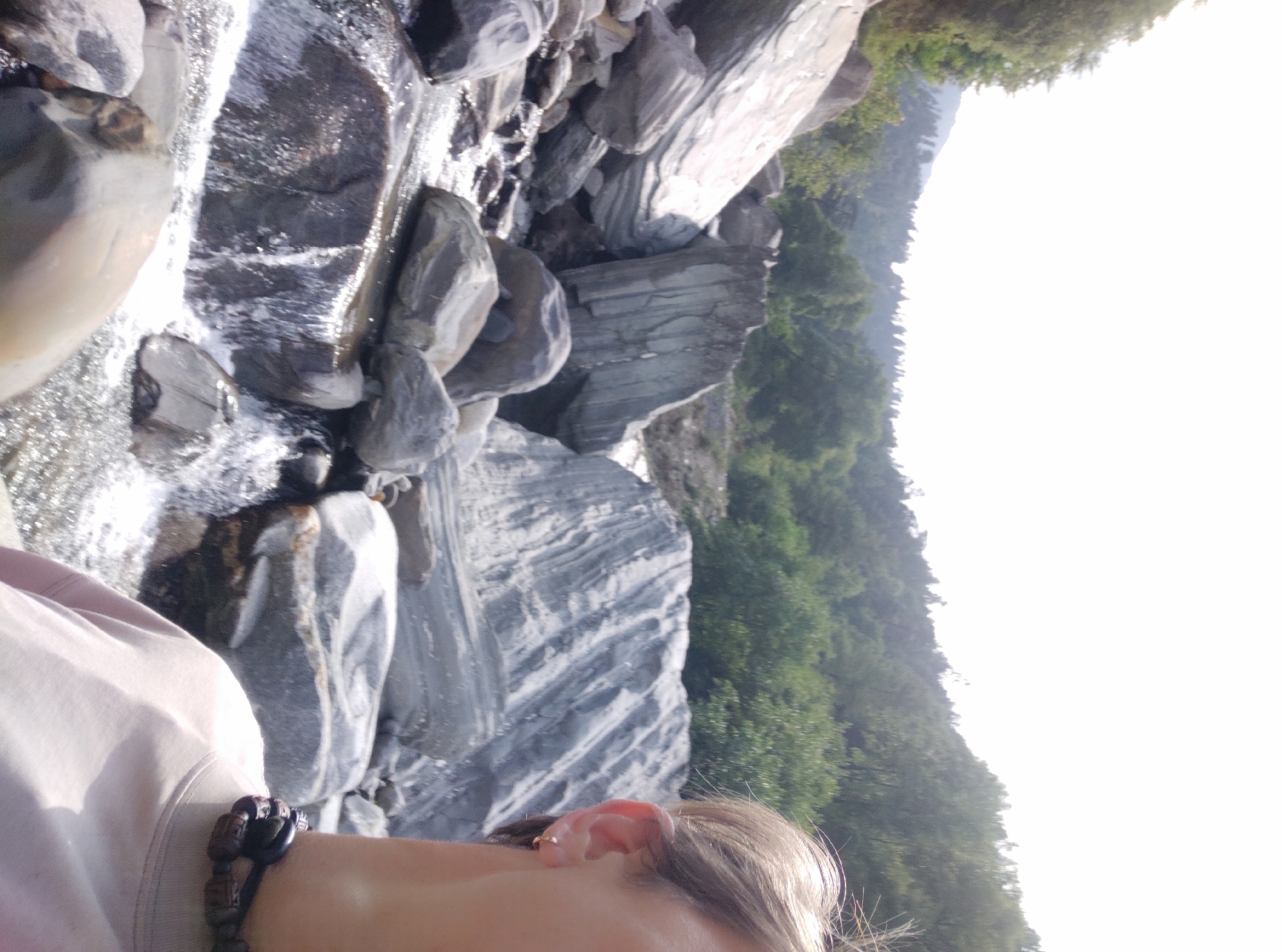 An over-the-shoulder shot captures a person's profile from the right side of the frame, facing left. The person has light-colored hair, an ear with a small gold hoop earring, and is wearing a light-colored t-shirt and a dark beaded necklace. The background is a natural landscape featuring a rocky riverbed with flowing water. Large, smooth, grey rocks are scattered throughout the scene, some of which have distinct striations. Beyond the riverbed, a lush green forested hillside rises, with the sky a bright, washed-out white at the top of the frame. The scene appears to be a tranquil outdoor location, possibly in the city of Baragran, India, given the natural elements.