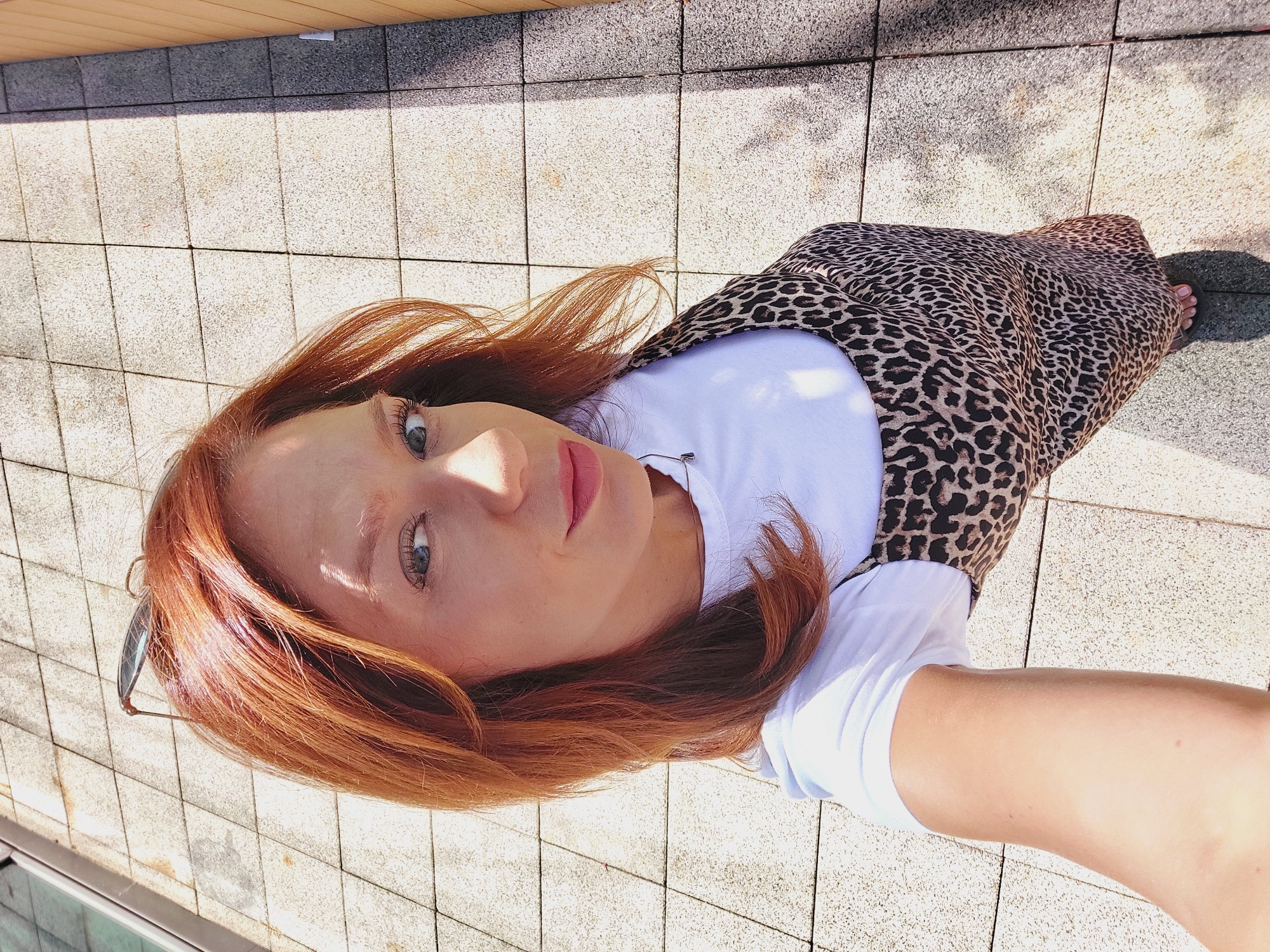 A young woman with reddish-brown hair and blue eyes is taking a selfie outdoors. She is wearing a white t-shirt under a leopard print dress and sandals. She is standing on a tiled patio with a wooden wall in the background. The image was taken in Sofia, Bulgaria.