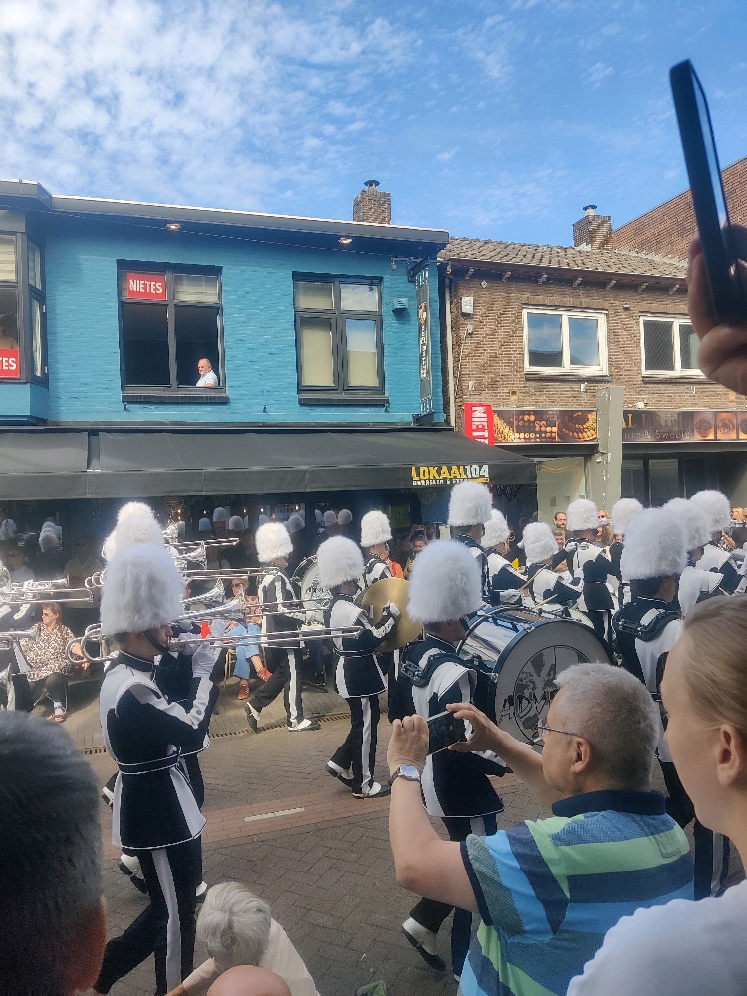 In Zundert, Netherlands, a marching band with members in black and white uniforms, adorned with tall white feathered hats, proceeds down a street. They are playing brass instruments and carrying drums. Spectators line the street, some sitting and others standing, observing the parade. One man in the foreground is capturing the event with his smartphone. Behind the band, buildings with storefronts and windows are visible, including a blue building with a sign that reads "NIETES" in red letters. The sky above is blue with scattered clouds. The ground is paved with cobblestones.