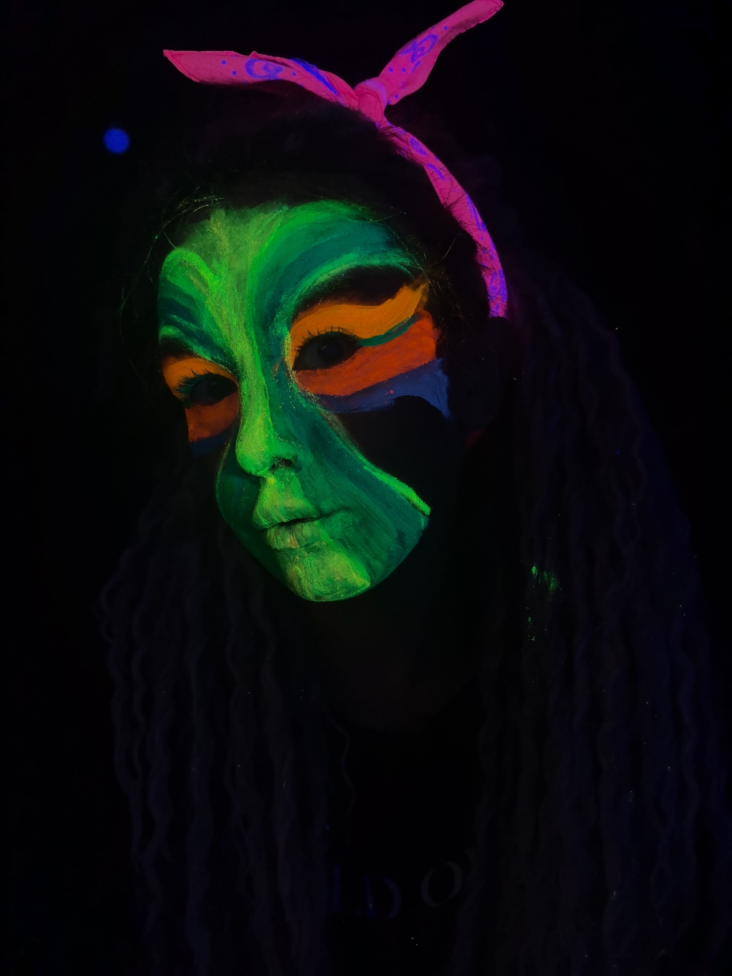 A person with dreadlocks is wearing neon face paint and a pink headscarf. The face paint is primarily green around the face with orange around the eyes and black around the forehead. The image is taken in a dark environment and appears to be lit with black light. The location is not discernible and does not appear to be in the city of Noviy, Russia.