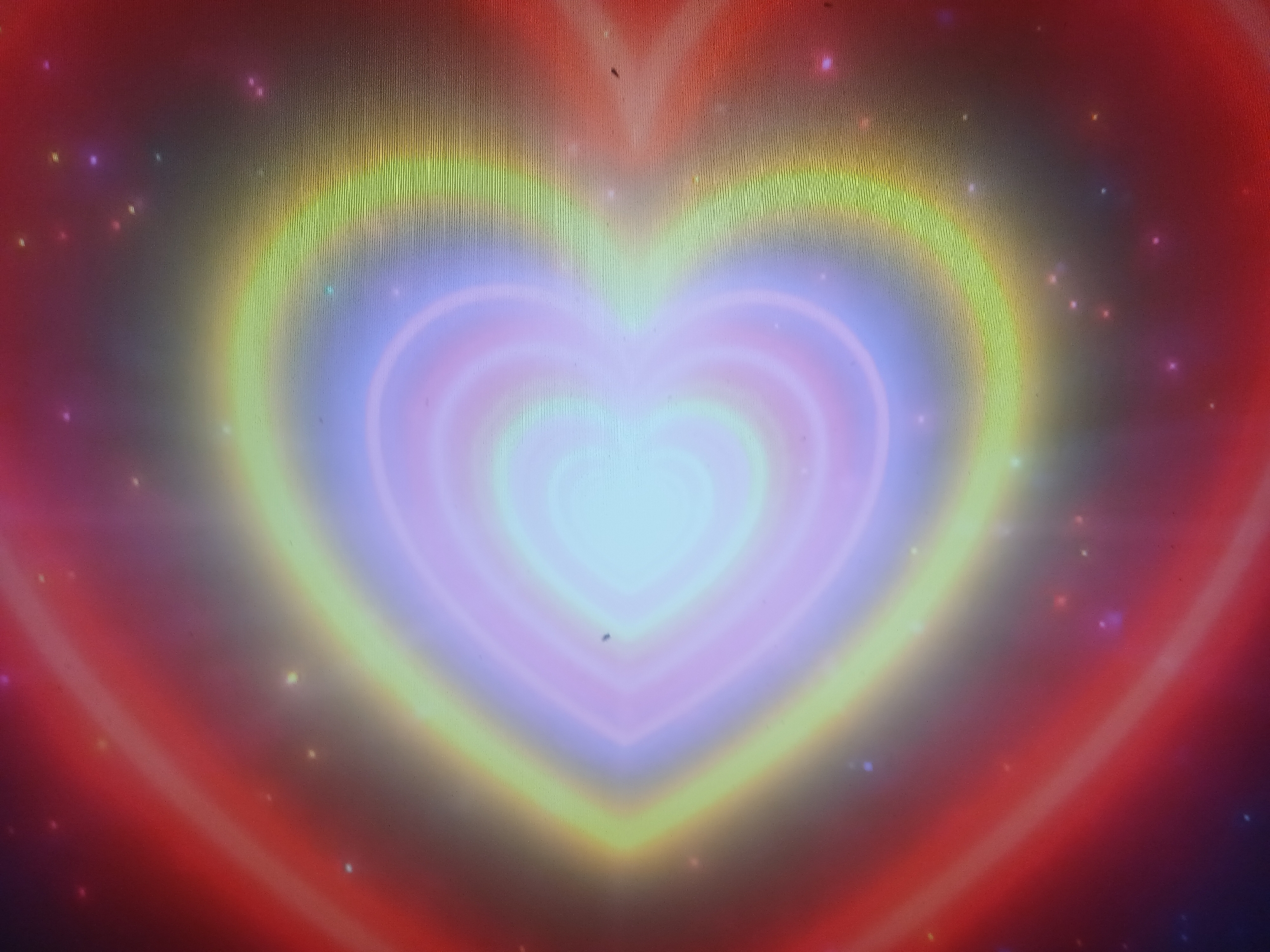 This is a close-up, abstract image featuring concentric heart shapes radiating from the center. The colors transition from a bright, pale blue or white at the core to soft pinks, purples, and yellows, then to a warm red glow at the outer edges. Scattered throughout the image are small, colorful bokeh-like lights that resemble stars or glitter. The overall impression is ethereal and vibrant, evoking a sense of warmth, love, or a cosmic phenomenon. There is no identifiable location in Parkent, Uzbekistan, or any discernible people, objects, actions, or interactions. The image is purely a stylized visual representation.