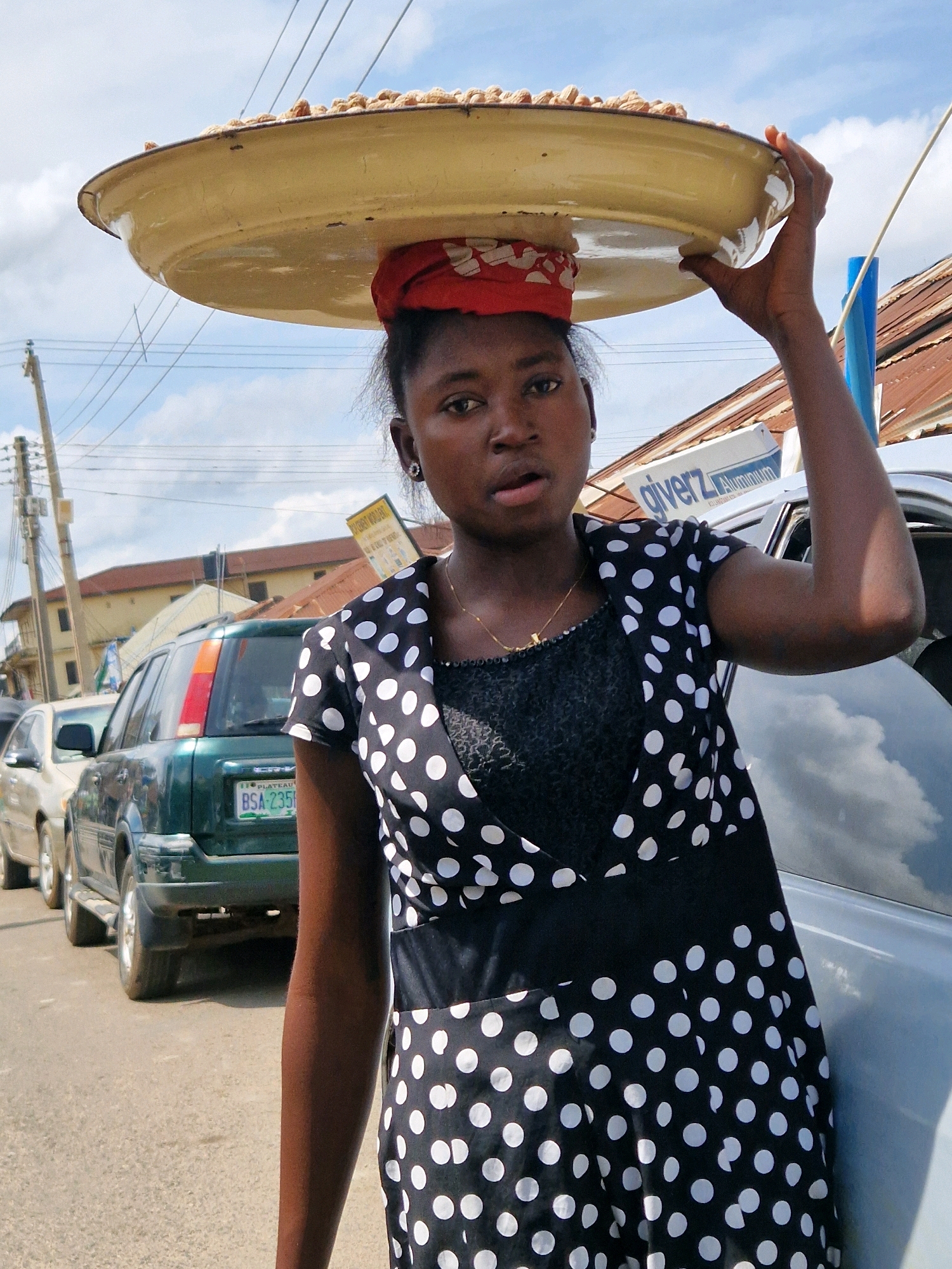 In the city of Jos, Nigeria, a young woman with dark skin carries a large, shallow, yellow tray on her head, laden with peanuts. She wears a red and white patterned headscarf and a black dress with white polka dots. She is standing on a paved road, with cars parked behind her. One car is green and has a license plate that reads "PLATEAU BSA-235H". The other cars are partially visible. In the background, buildings with corrugated metal roofs are visible, as are utility poles and overhead wires. The sky is partly cloudy.