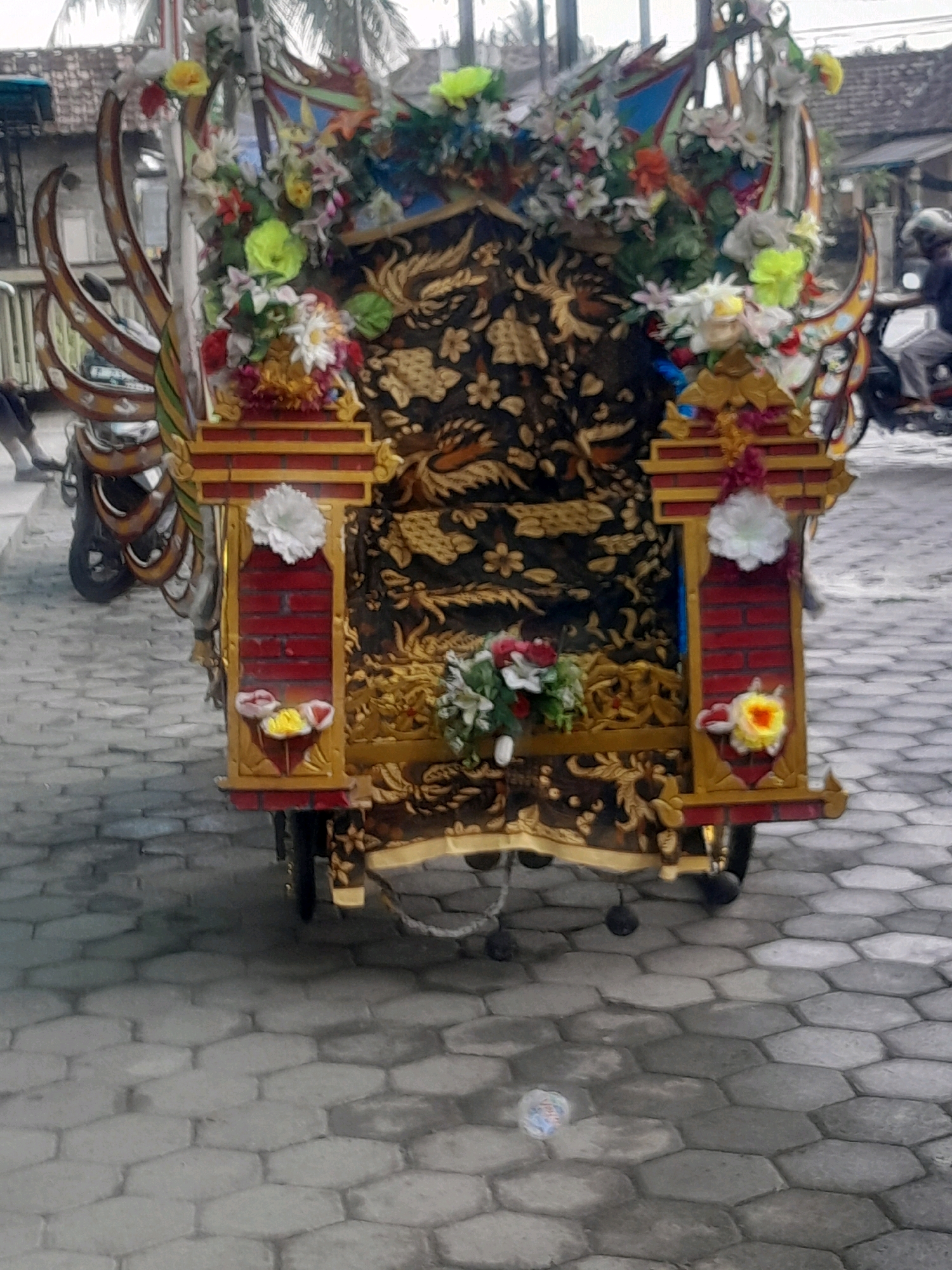 A horse-drawn carriage, adorned with flowers and intricate golden patterns, is parked on a cobblestone street. The carriage has a dark, ornate fabric with a golden dragon motif. Red and gold decorative elements are visible on the sides, along with white and yellow flowers. A person on a motorcycle is visible in the background to the right, wearing a helmet. The street appears to be in a city, with buildings visible in the background.