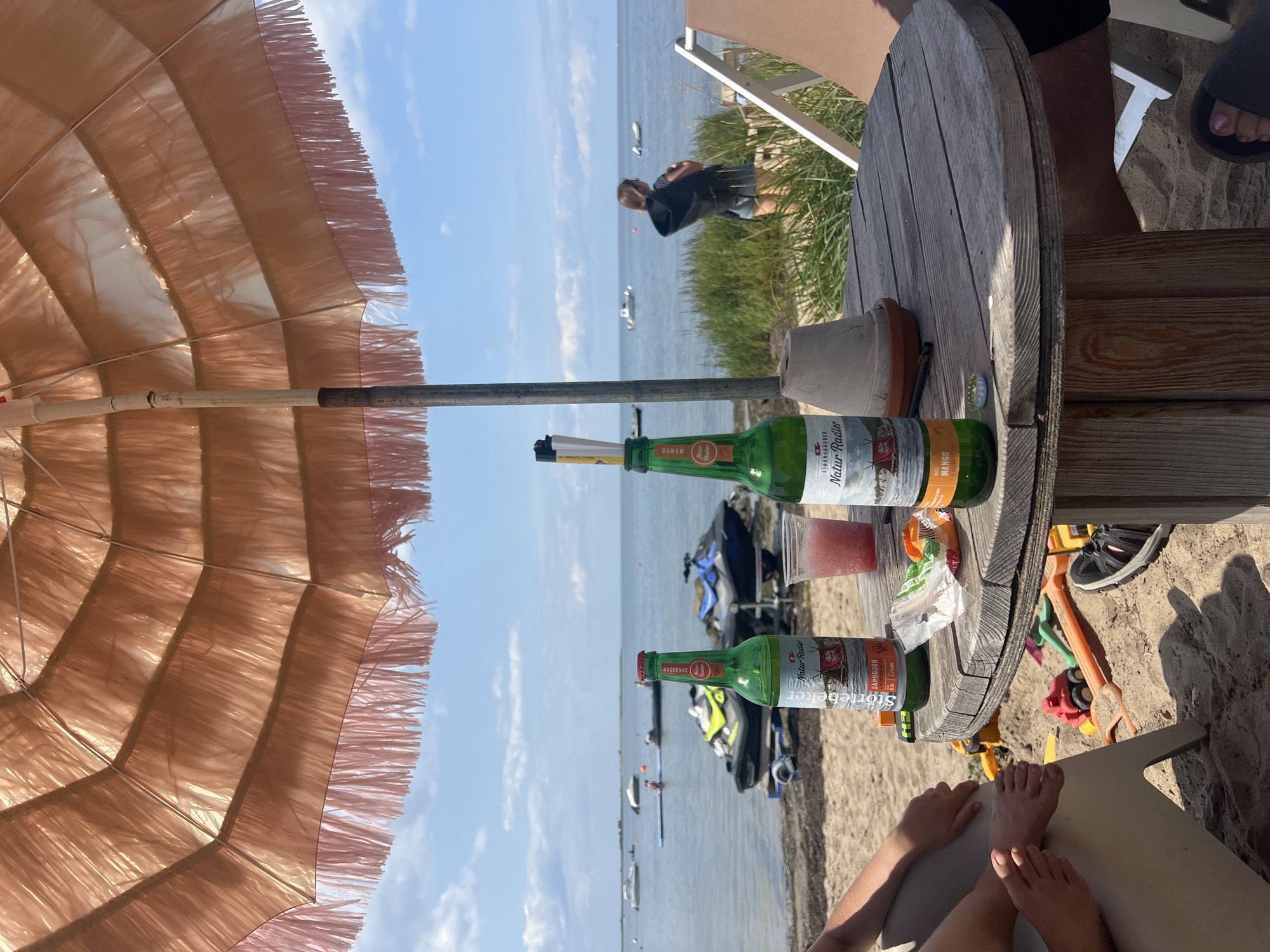 The image depicts a scene on the beach at the Baltic Sea coast near Boiensdorf, Germany, under a pale pink fringed parasol. A round, weathered wooden cable spool serves as a table, on which sit two green glass bottles of Natur Radler beer (one labeled "Storckbeeren" and the other "Mango"), a plastic cup containing a pink frozen beverage, a small grey pot, and some wrappers. In the foreground, a person's legs and feet are visible resting on a beige lounge chair. Scattered in the sand near the table are children's beach toys, including a red toy truck and an orange shovel. The background shows the calm sea with several boats and jet skis, and a woman in the distance looking out at the water. The sky is blue with scattered clouds.
