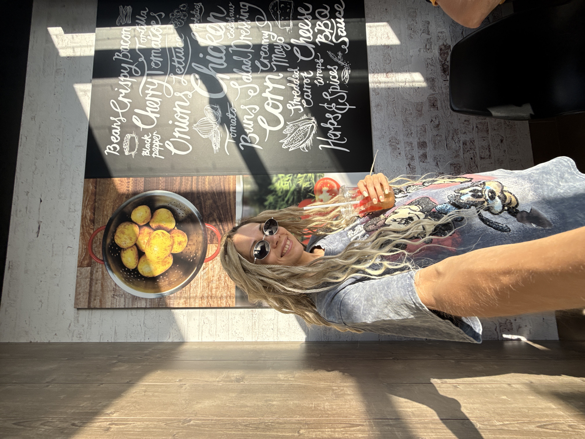 A young adult woman with long blonde hair, wearing sunglasses and a grey t-shirt with a Mickey Mouse print, smiles at the camera while holding a red drink. She is standing in front of a wall with a large menu board that lists various food items in white chalk lettering, including "Beans," "Crispy Bacon," "Cherry Tomatoes," "Onions," "Chicken," "Corn," "Cheese," and "BBQ Sauce." There is also a framed image of potatoes in a colander on the wall. The environment appears to be a restaurant, with wooden wall paneling and brickwork visible. Sunlight streams in from the side, casting shadows across the scene. The location is stated to be Шебунино, Russia.