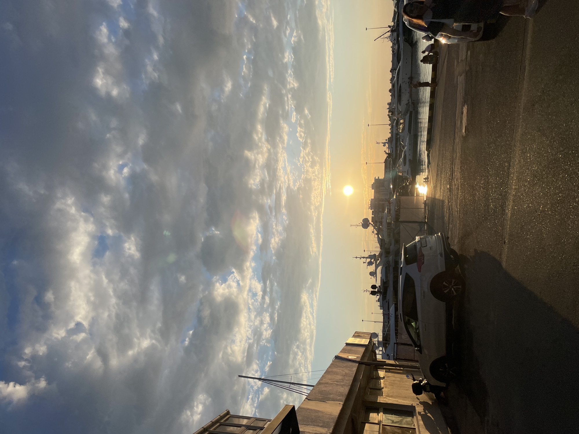 This media file captures a scenic view of a harbor at sunset, likely in Sochi, Russia. The sun, low on the horizon, casts a warm, golden glow across the sky, illuminating a scattering of dramatic clouds. The sky transitions from a bright, sunlit expanse to darker, more ominous storm clouds on the left.
In the foreground, a street or pathway runs alongside the harbor. A white SUV is parked on the right side of the image, its shadow stretching long across the wet asphalt, suggesting recent rain or mist. Further down the pathway, parts of buildings and structures are visible, hinting at a developed port area.
The harbor itself is filled with various yachts and boats, docked along the waterfront. The water reflects the warm light of the setting sun. A few people are visible, including individuals standing near the parked car and on a walkway. The overall atmosphere suggests a peaceful end to the day, with a touch of dramatic flair from the contrasting sky conditions. The time of day is clearly sunset, and the weather appears to be post-rain, with a pleasant, soft light. No visible text is present in the image.