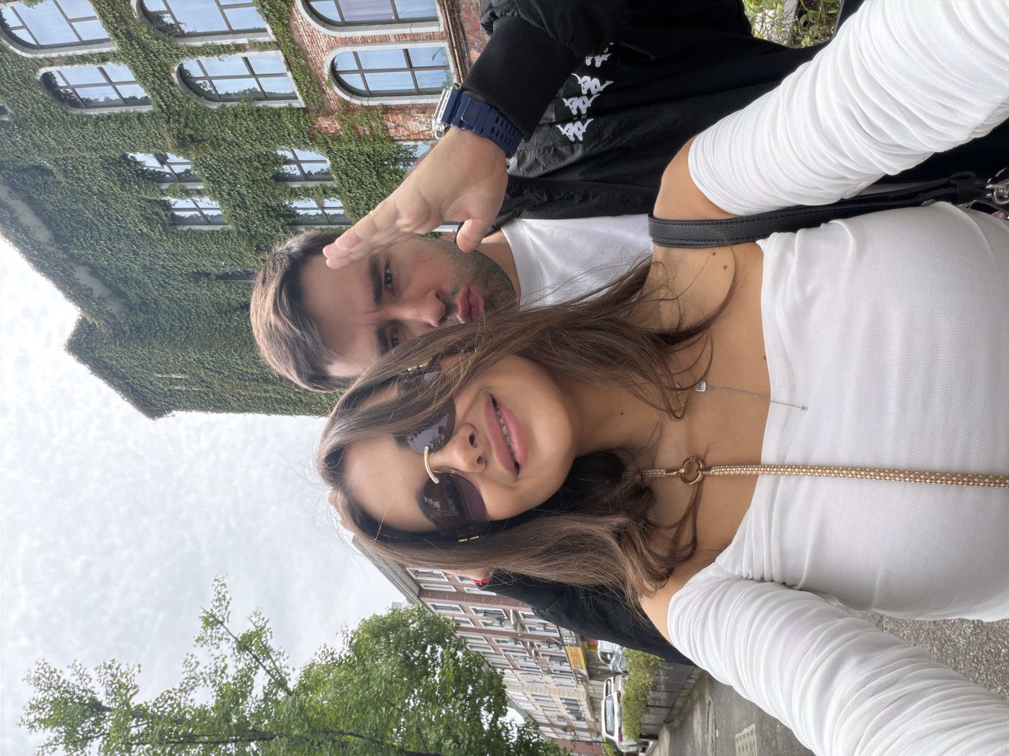 A man and a woman are posing for a selfie outdoors, likely in an urban area. The woman is in the foreground, wearing sunglasses, a white top with long, gathered sleeves, and necklaces. The man is behind her, wearing a black jacket over a white shirt and a blue watch. His hand is raised in a casual gesture. Behind them are buildings; one is covered in green ivy, and another is made of red brick. The sky is overcast. No clear indications suggest the location is Zhangjiajie, China.