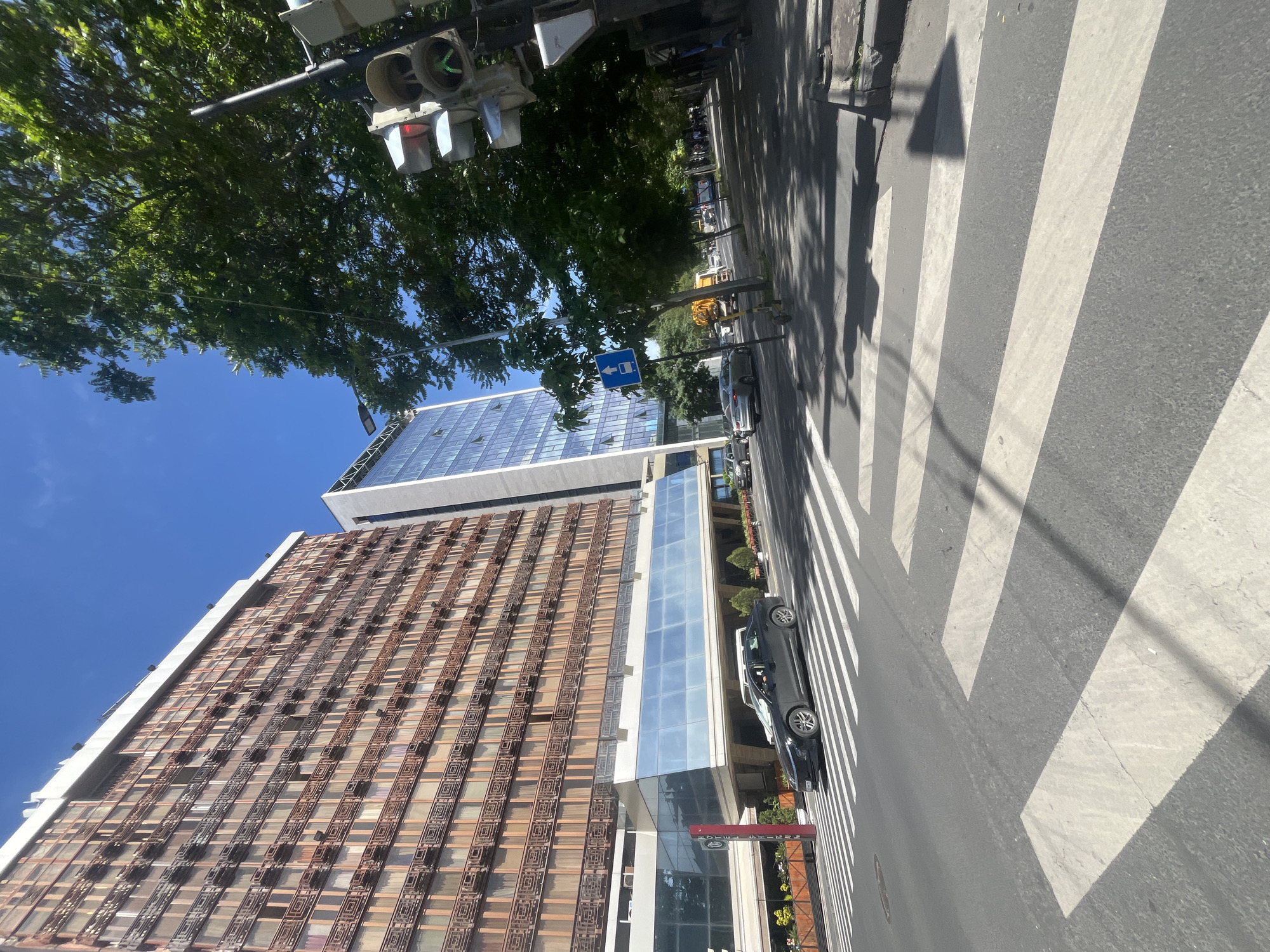 The image depicts a street scene in Yerevan, Armenia. A crosswalk with white stripes stretches across a paved road. A black car is parked on the road near the crosswalk. A building with a facade featuring a grid-like pattern is visible on the left side of the frame. Adjacent to this building is another, taller, glass-facaded building. Overhead, traffic lights are suspended above the street, with visible greenery from trees lining the road. The sky is a clear blue. A blue bus stop sign is positioned further down the street. The scene is captured in daylight.