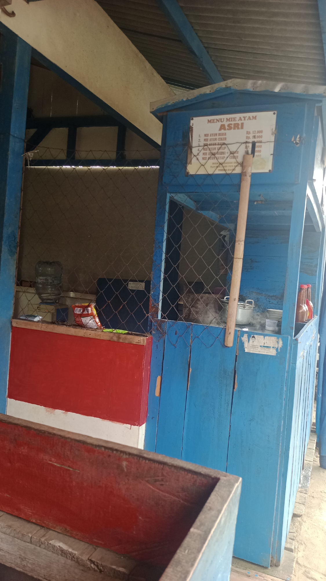 The image shows a small, open-air food stall painted bright blue and red, located on a sandy path in Cikalong, Indonesia. A sign above the counter lists "MENU MIE AYAM ASRI" with prices for various noodle dishes. Inside the stall, a water dispenser and a bag of snacks are visible on a shelf. In the background, a metal fence encloses the preparation area, where cooking pots and bottles of condiments are present. The stall appears to be situated near a building with a corrugated metal roof. A wooden bench, also painted red, is in the foreground. The overall impression is of a humble, local eatery.