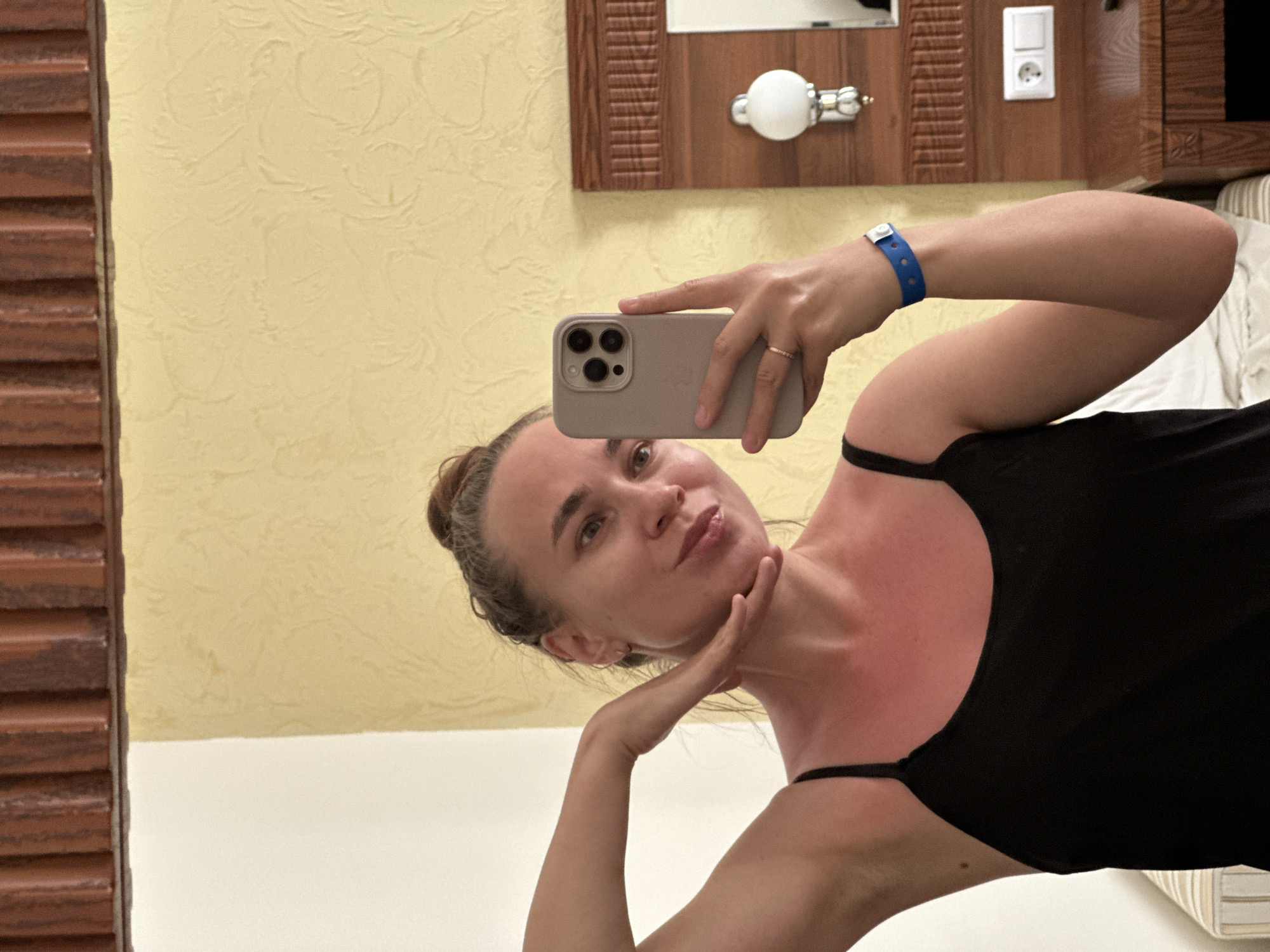 The media file is a selfie taken by a young woman in a hotel room in Vityazevo, Russia. The woman has her hair pulled back in a bun and is wearing a black tank top. She is holding a smartphone in her left hand, taking the picture in a mirror. Her right hand is touching her chin. She has a blue wristband on her left wrist. The background shows a yellow textured wall, a wooden headboard with a light fixture and an electrical outlet, and a section of a striped curtain. The overall lighting suggests it is daytime. The woman appears to be posing playfully for the photo, with a slight smile and puckered lips. The room appears to be a hotel room, as indicated by the décor and the presence of a wristband, possibly for an all-inclusive resort.