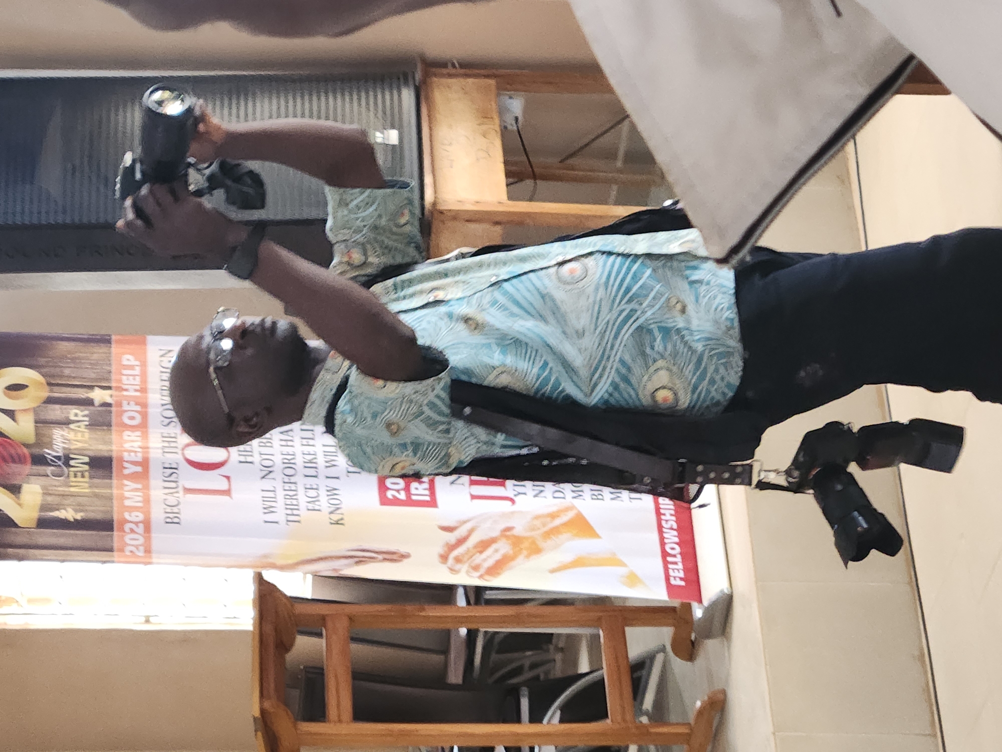 The image depicts an indoor scene, possibly a church or a community hall, with a banner in the background that reads "2026 MY YEAR OF HELP". A man, appearing to be of African descent, is the central subject. He is wearing glasses and a light blue shirt with a peacock feather pattern, and dark trousers. He is holding a camera up to his eye, as if taking a photograph, and another camera is hanging from a strap around his neck. The background includes a wooden podium or shelf, and part of a wooden table with some writing on it. The lighting suggests it is daytime, possibly with natural light coming from a window off-frame. The overall impression is of an event or gathering where photography is taking place. The presence of the banner with a specific year and a religious-sounding phrase suggests a context of faith or community. The city of Jos, Nigeria, is mentioned in the prompt, but there are no specific visual cues in the image that definitively place it in Jos.