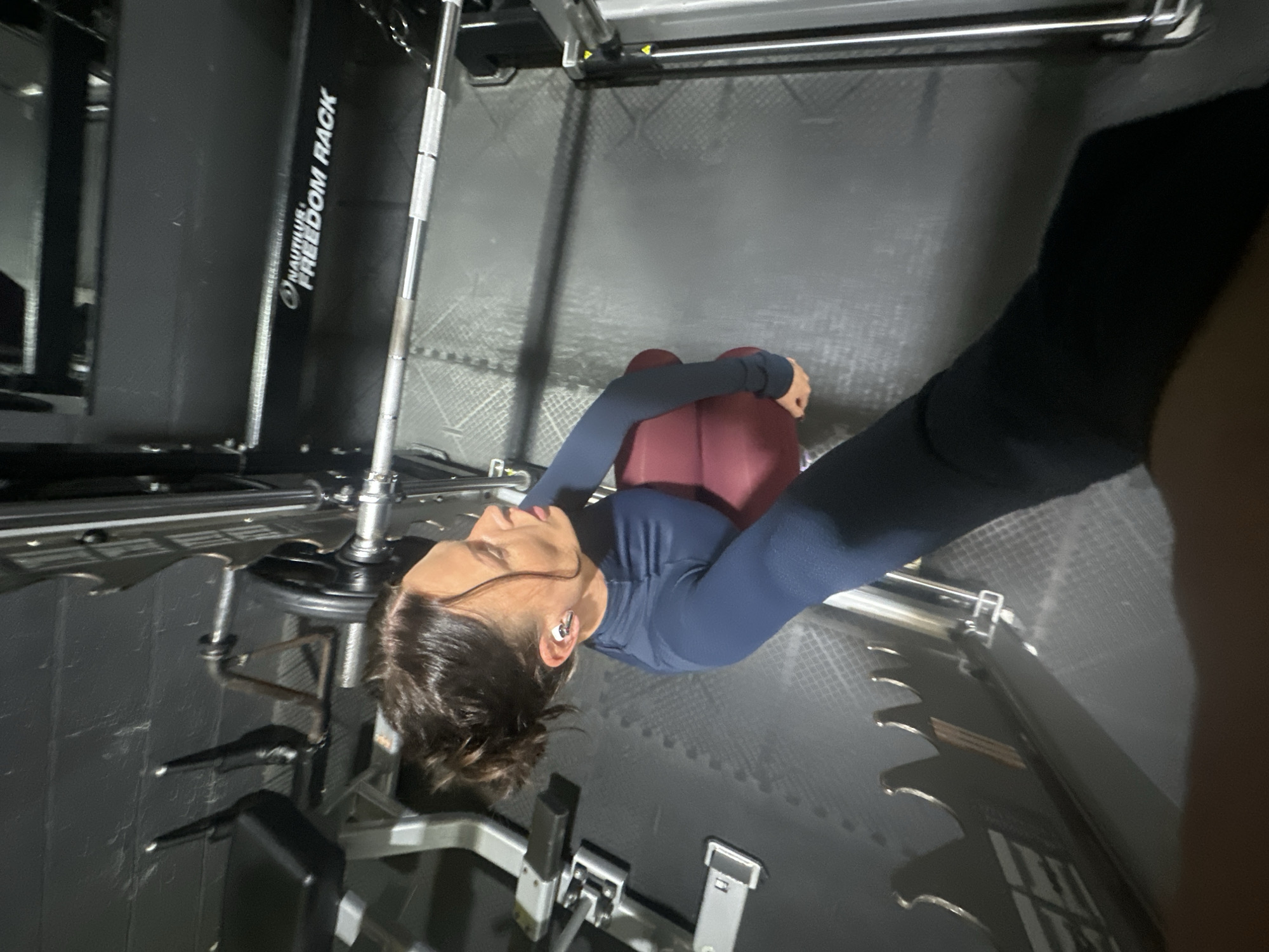 A person wearing a long-sleeved blue shirt and maroon pants is sitting on the floor of a gym in Tonbridge and Malling, United Kingdom. They have short brown hair in a bun and are wearing an earbud. The gym contains workout equipment, including a Nautilus Freedom Rack. The floor is a grey rubber mat.