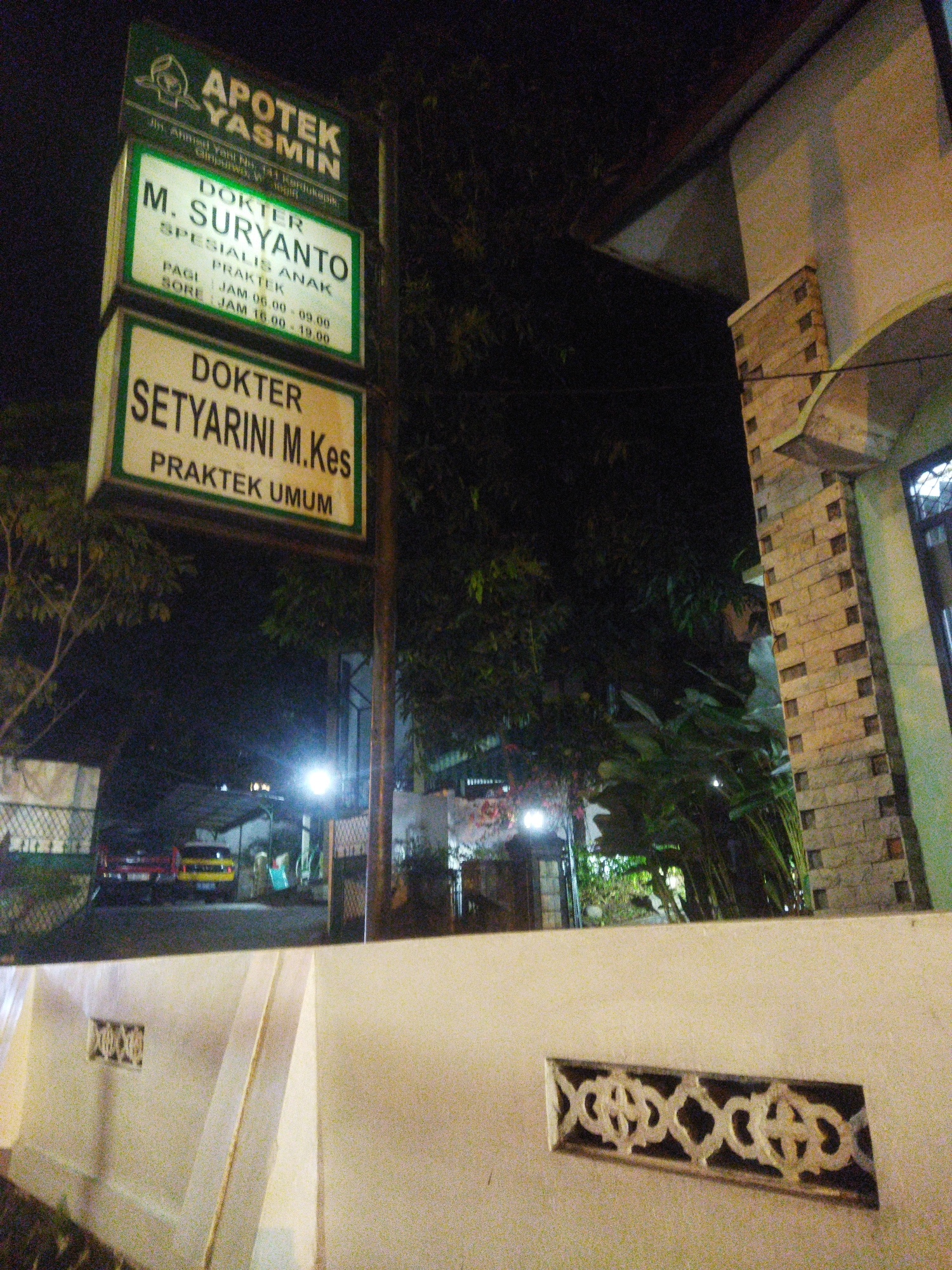 The image shows a sign for "APOTEK YASMIN" which is a pharmacy. Below the pharmacy sign, there are two signs for doctors. The first sign indicates "DOKTER M. SURYANTO" who is a specialist for children and a practitioner. His practice hours are from 06:00 to 09:00 in the morning and 16:00 to 19:00 in the afternoon. The second sign indicates "DOKTER SETYARINI M.Kes" who is a general practitioner. The signs are located in front of a building, likely a clinic or pharmacy, at night. The background shows trees, a street with a yellow car and a red car, and buildings in the distance. A wall with decorative patterns is in the foreground. The location is specified as being in the city of Wonogiri, Indonesia.