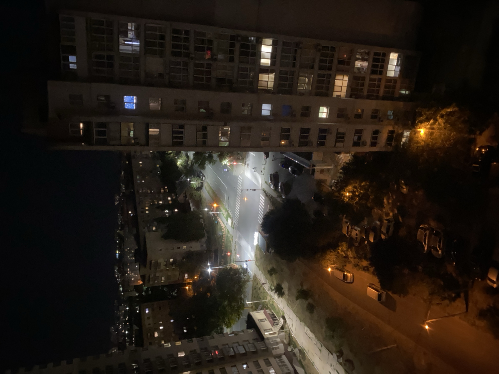 An exterior, high-angle, night-time shot captures an urban scene in Chornomorsk, Ukraine. The primary focus is on a tall, multi-story residential building with numerous lit windows, indicating occupancy. To the left of this building, another similar structure is partially visible. A street intersection with pedestrian crosswalks is visible, illuminated by streetlights. Several parked cars are present along the side of the road and in parking areas. Trees and other foliage are scattered throughout the scene, adding greenery to the urban landscape. The sky is dark, typical of nighttime.
