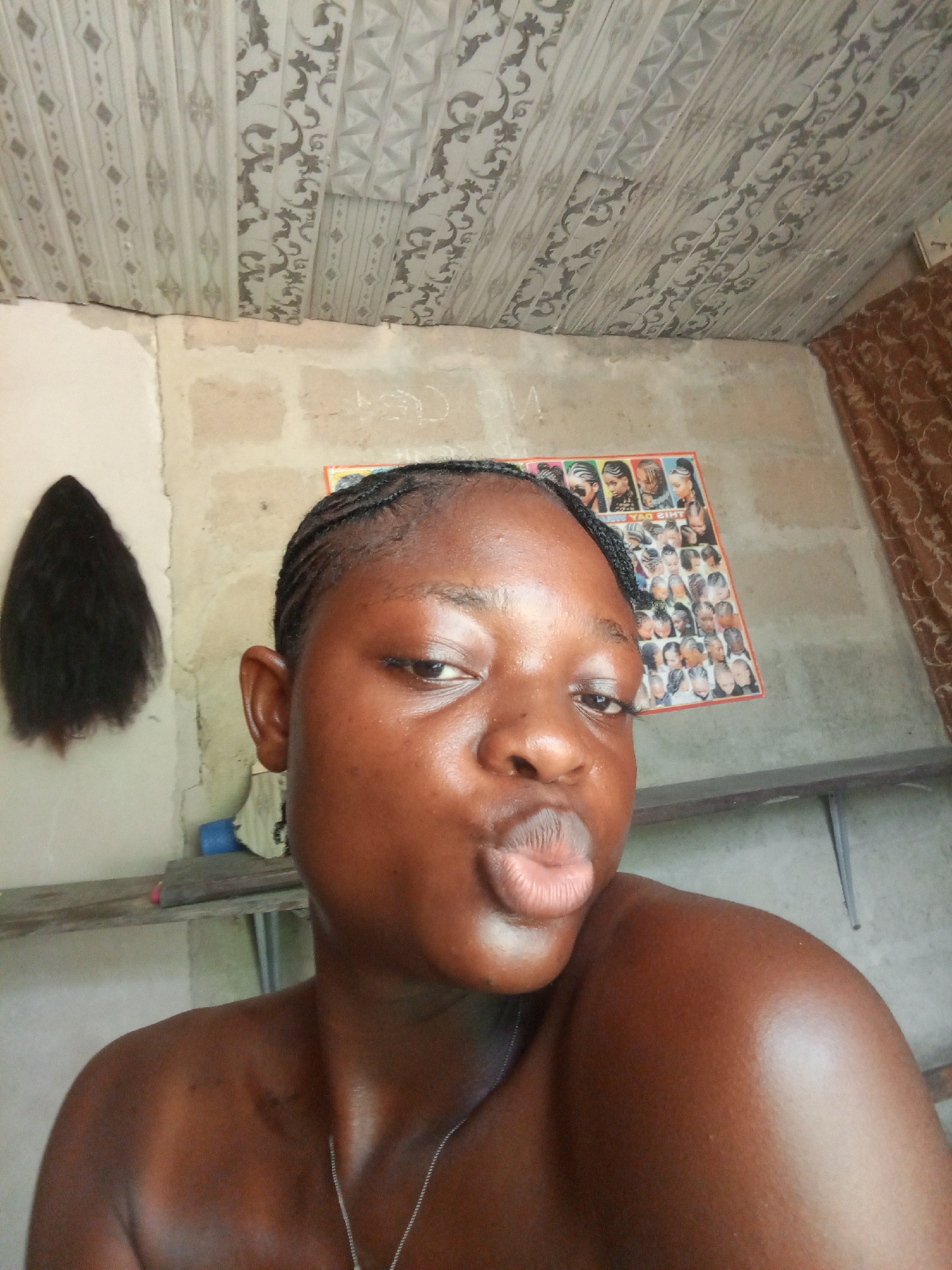 A close-up, eye-level indoor shot shows a dark-skinned woman with braided hair, puckered lips, and a silver necklace, posing for the camera. Behind her, the wall is unfinished, with a poster showing various hairstyles attached to it. A dark-haired wig hangs on the wall to the left. There is a wooden shelf with objects on it near the wall. The ceiling is tiled with a patterned design, and a curtain hangs partially visible on the right side of the wall. The location is likely Uyo, Nigeria.