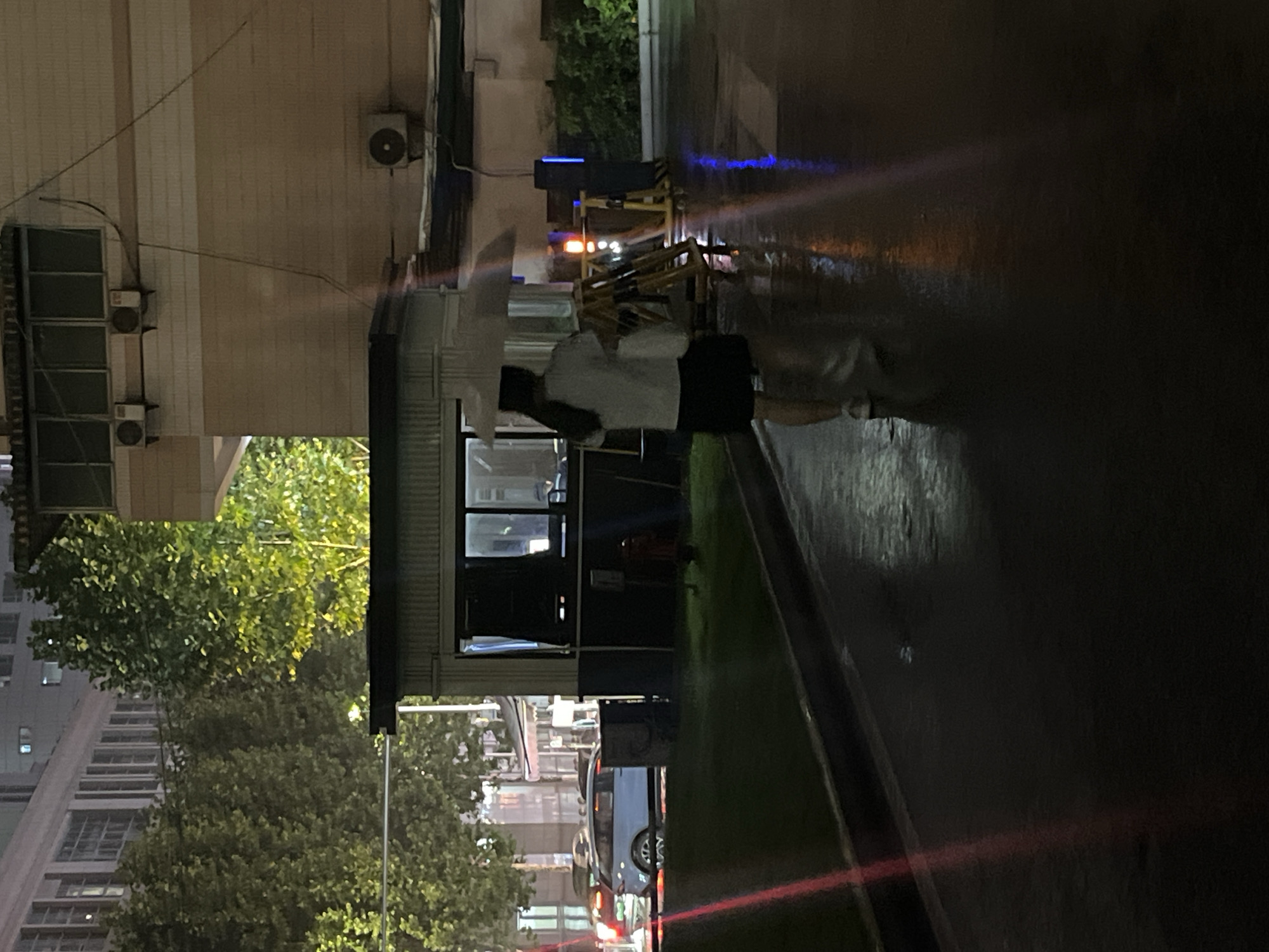 The image captures an urban outdoor scene at night within Yuzhong District, China, characterized by wet, reflective surfaces indicative of recent rain. In the foreground, a single individual, viewed from behind, walks away from the camera on a dark, paved pathway. This person, wearing a light-colored short-sleeved top and a dark skirt or shorts, holds a white umbrella overhead and carries a light-colored bag in their left hand.
To the left of the individual, a compact, dark-colored guard booth or security structure with illuminated windows stands prominently. Behind and to the left of this booth, a light-colored vehicle with illuminated taillights is partially visible on an adjacent road. The background features several multi-story buildings partially obscured by dense green trees, which are brightly illuminated by artificial lights.
On the right side of the paved pathway, a configuration of yellow and black striped barriers or tripods is present, accompanied by an active orange warning light and a tall, blue illuminated pole-like device. The wet ground reflects these light sources in elongated patterns. Further right, a multi-story building with a light-colored facade, windows, and external air conditioning units is visible. A red fire hydrant is positioned on a grassy strip between the paved path and the guard booth. Overhead utility wires traverse the upper portion of the frame. A distinct red light streak, consistent with lens flare, extends diagonally from the lower left towards the upper right through the center of the image.