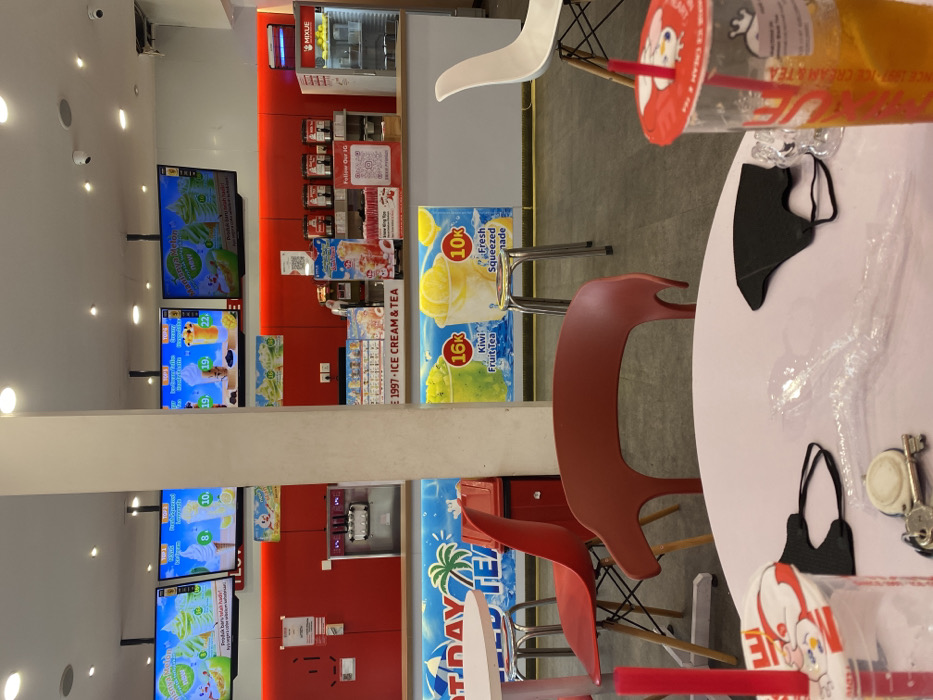 The image shows a cafe interior with a white table and a black face mask on it. Two red chairs are in front of the table. A glass of orange liquid with a red lid and a straw is in the foreground. Behind the table, there is a counter with a red-and-white sign reading "1997-ICE CREAM & TEA". On the counter are displays featuring "Kiwi Fruit Tea" for 16k and "Fresh Squeezed" for 10k. Two red chairs are facing the counter. The walls are red with advertising displays showing images of various drinks. The location is in Kecamatan Piyungan, Indonesia.