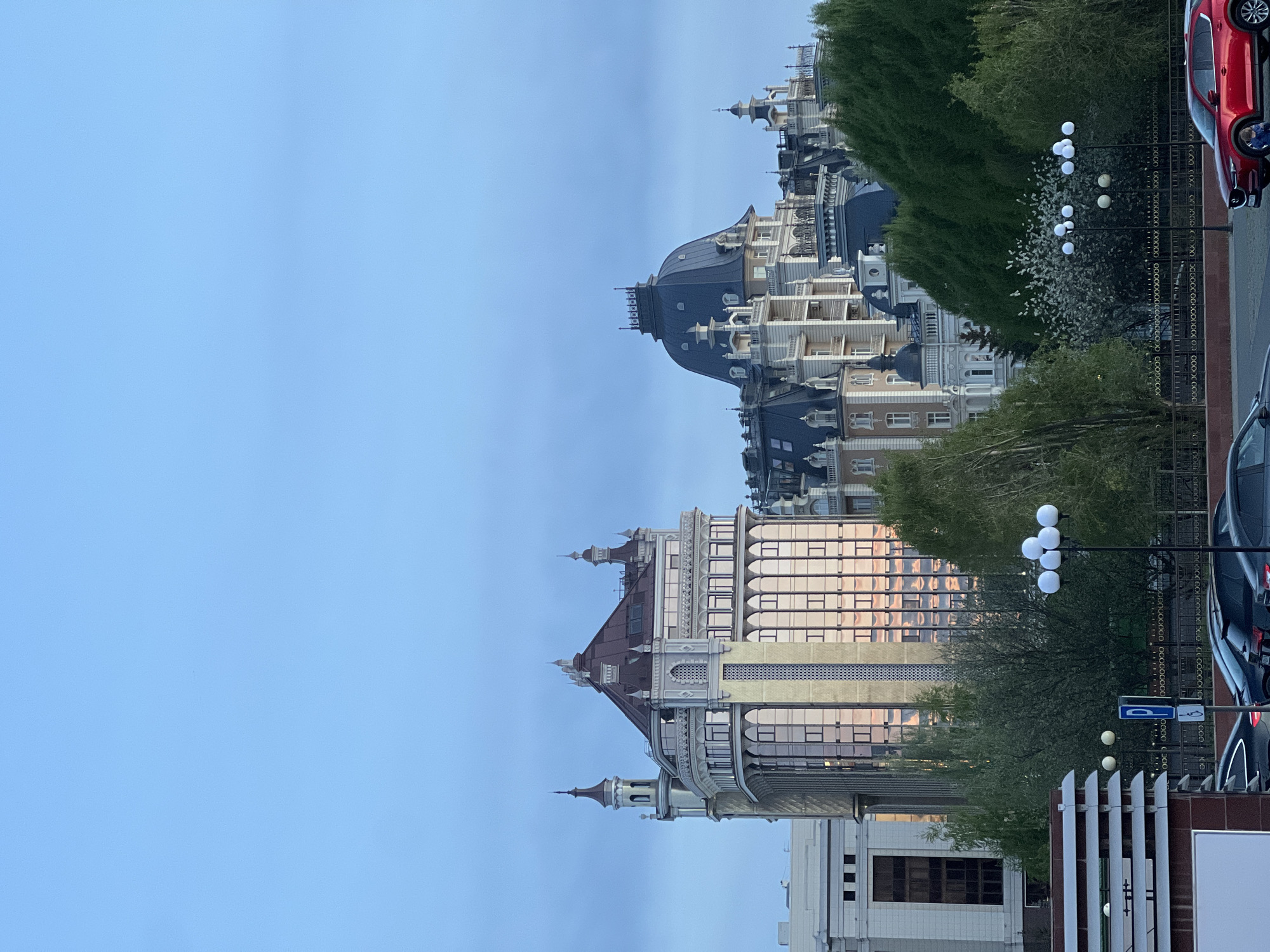 The image depicts a cityscape in Дачное, Russia, featuring ornate buildings with European architectural styles. The foreground includes a park area with trees, decorative lampposts with spherical lights, and a wrought-iron fence. Beyond the fence, a parking area contains parked cars. The buildings showcase a variety of architectural elements, including domes, towers, and multiple levels with detailed facades. The sky is a clear, light blue, suggesting either early morning or late afternoon lighting.