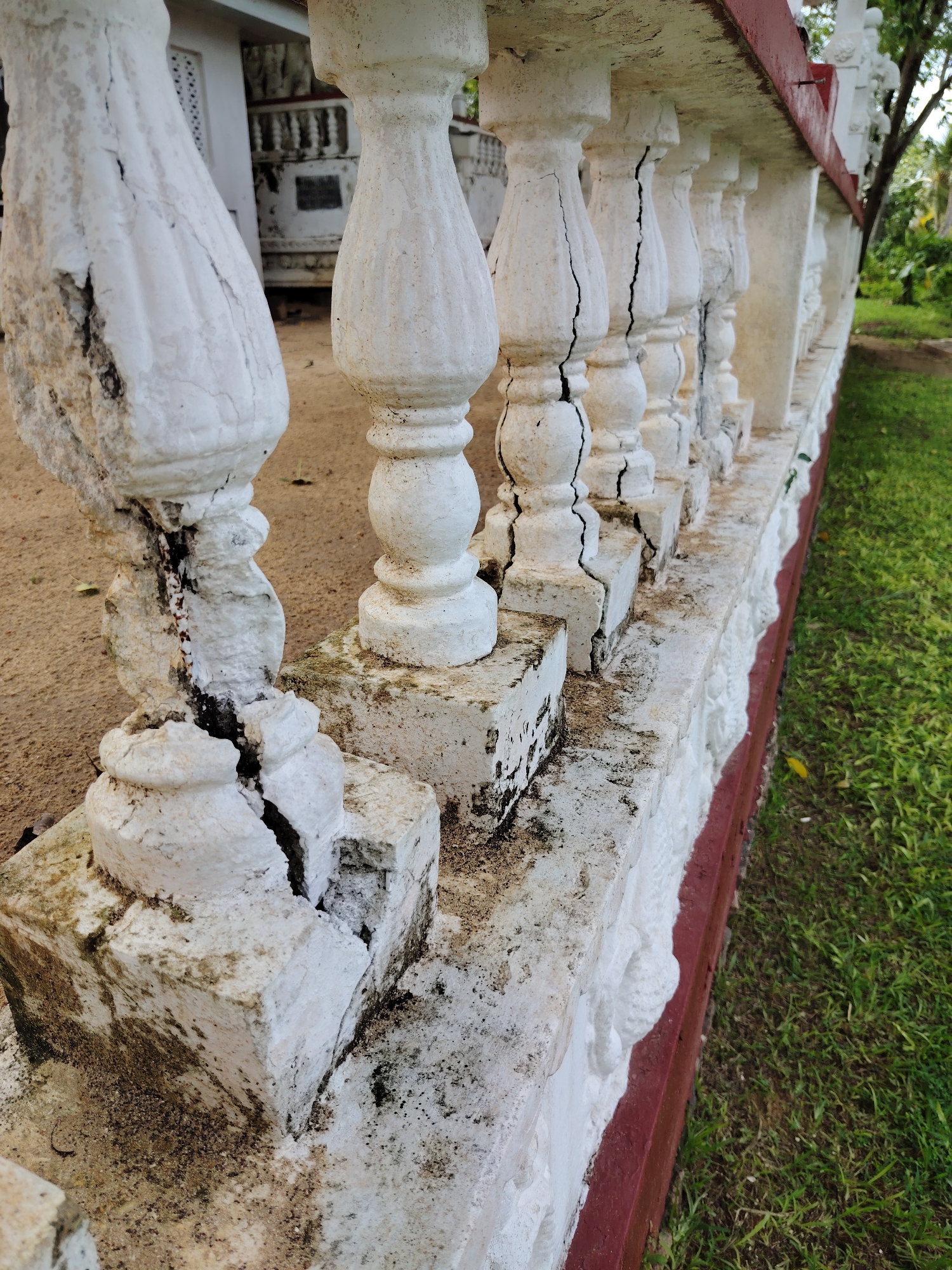 The image displays a close-up, angled view of a deteriorated white balustrade or railing. A row of white, urn-shaped balusters, made of what appears to be concrete or stone, runs diagonally across the frame from left to right. Many of these balusters exhibit severe vertical cracks, some extending from top to bottom, revealing darker material within their fissures. The white paint on the balusters and their supporting base is chipped, peeling, and discolored with dark patches of mold or dirt.
The balusters are anchored to a low, white wall or base that also shows signs of weathering and grime. Along the bottom edge of this white base, there is a prominent, dark red trim. Below this red trim, a strip of light green grass is visible on the right side of the frame. On the left side, behind the railing, the ground consists of light brown soil or sand. In the distant background, parts of a white building or structure with a dark window or opening are faintly visible. The lighting suggests it is daytime. There are no people or discernible activities taking place, and no visible text.