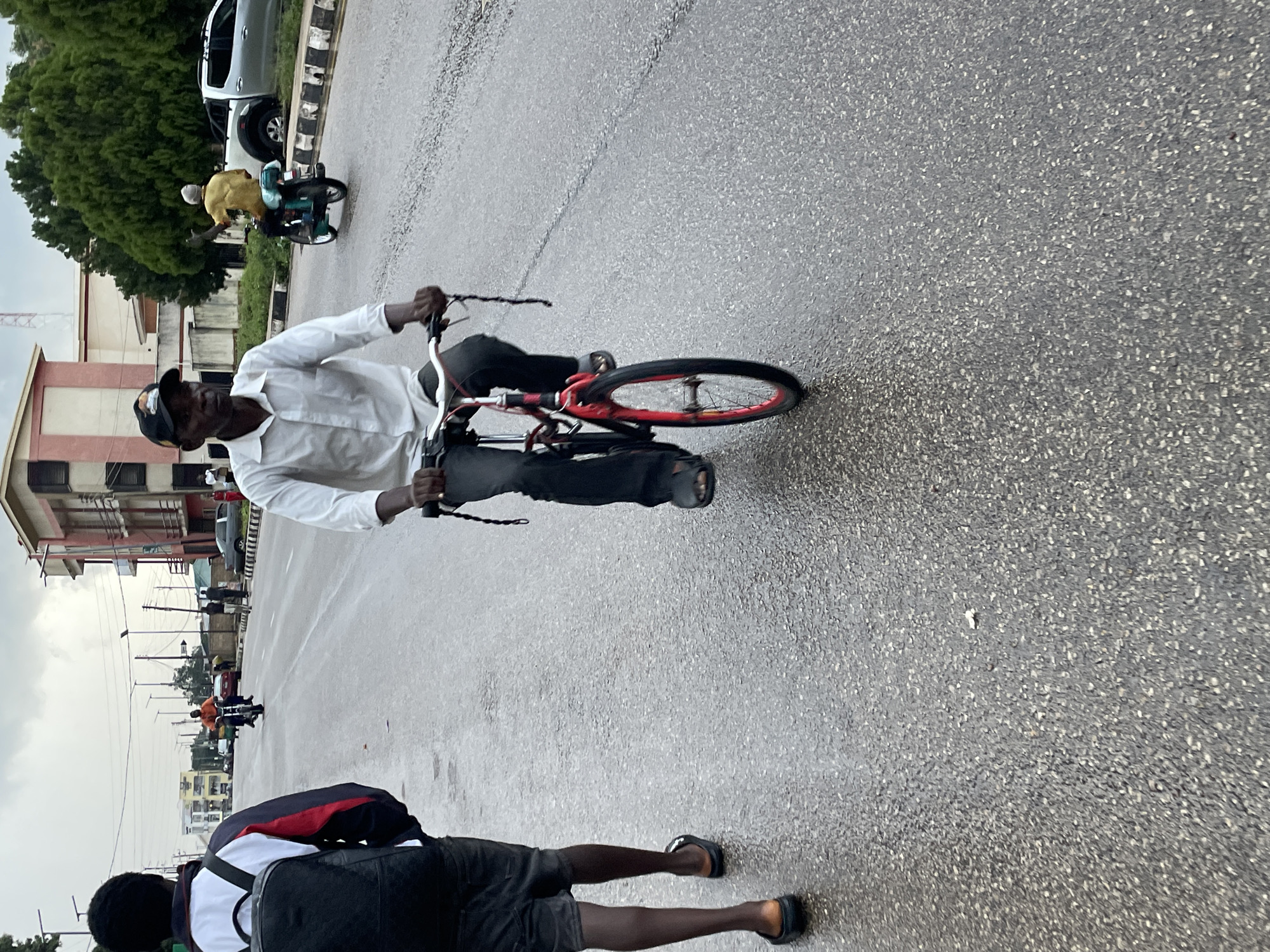 Two young Black men are walking on a wet, grey asphalt road in Jos, Nigeria. The man on the left is wearing a black backpack with white and red accents, dark shorts, and black slides. The man on the right is on a red bicycle, wearing a white button-down shirt, black pants, and black sandals. He is looking forward as he pedals, and a dark braided cord hangs from the handlebars. The road is lined with buildings, some of which are multi-story with light-colored walls and dark roofs. Trees with dense green foliage are visible in the background. The sky is overcast, suggesting a cloudy or rainy day. Several other people and a motorbike are further down the road, indicating a somewhat busy urban environment. The image appears to have been taken from a low angle, close to the ground.