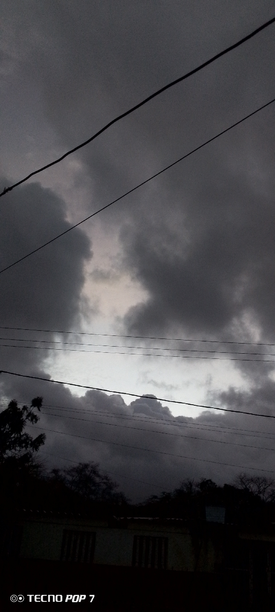 The image presents a dramatic, heavily overcast sky, filled with dense, dark grey clouds that suggest impending rain or a recent storm. Several black power lines traverse the frame horizontally, creating stark lines against the varying shades of the sky.
In the upper middle section, a brighter, lighter grey area of the sky breaks through the darker clouds, indicating a source of diffused light. A more prominent, elongated strip of bright sky runs across the mid-section of the image, sharply contrasting with the heavy cloud masses above and below it.
Towards the bottom left, the dark silhouettes of tree branches and foliage are visible. Below these, the top portion of a building with visible window frames can be seen, though it is largely dark and indistinct against the gloomy backdrop. The overall lighting is dim, characteristic of a heavily clouded day, late afternoon, or early evening.
The scene evokes a somber or contemplative mood due to the dark weather conditions. The setting is in Altagracia, Venezuela, as per the provided context. The bottom left corner features a watermark that reads "TECNO POP 7," identifying the camera used. No people or specific activities are visible in the image.