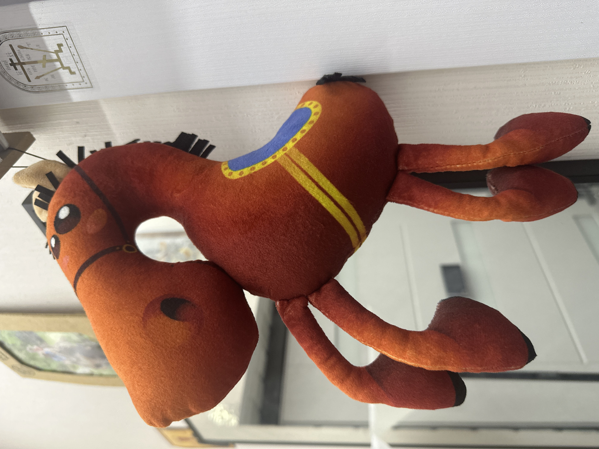 This media file features a plush toy horse with a saddle. The horse is primarily brown with black yarn for its mane. It has a blue and yellow patterned saddle and small white eyes with black pupils. The toy is positioned as if it is lying down.
In the background, the top portion of the image shows a white surface with a metallic emblem. The emblem appears to be a crest with a shield shape and intricate designs within it, including what looks like a stylized cross or figure. Below the emblem, some text is partially visible in black, but it is too blurry to read clearly. The overall setting seems to be indoors, possibly on a shelf or table, with the plush toy positioned in the foreground.