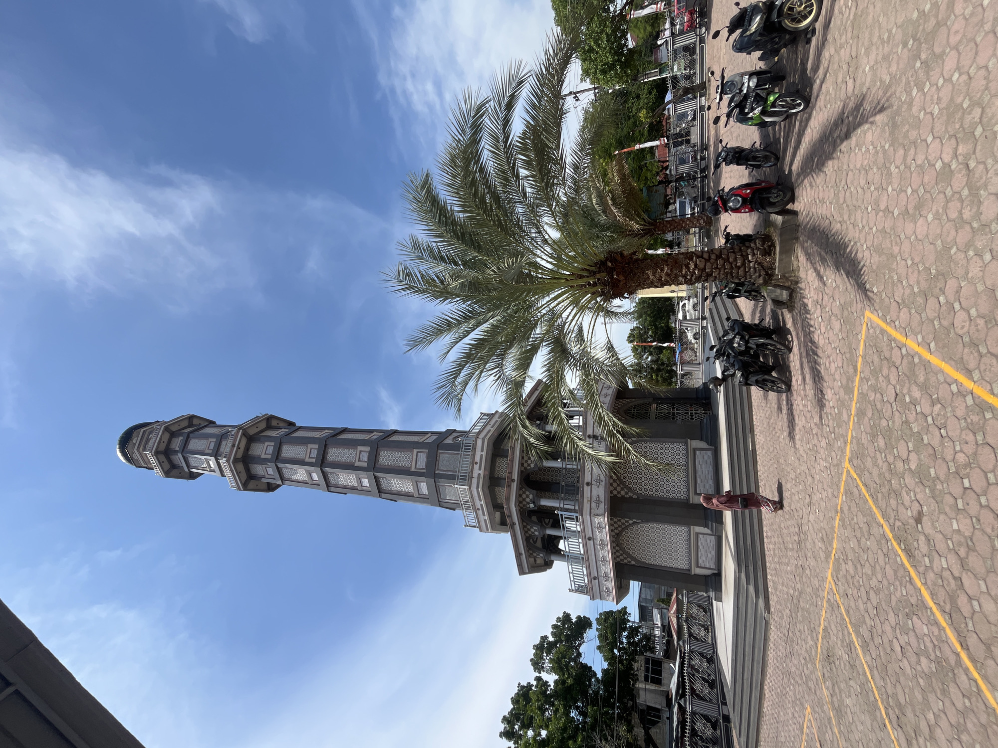 The image depicts a tall, gray tower under a blue sky with scattered clouds. The tower appears to be part of a mosque, displaying several stories with patterned windows and balconies. In the foreground, there is a palm tree next to the tower and a paved area, with motorcycles parked nearby. A person wearing a reddish-brown garment stands on the steps leading to the tower. The setting is in Keudekruenggeukueh, Indonesia.