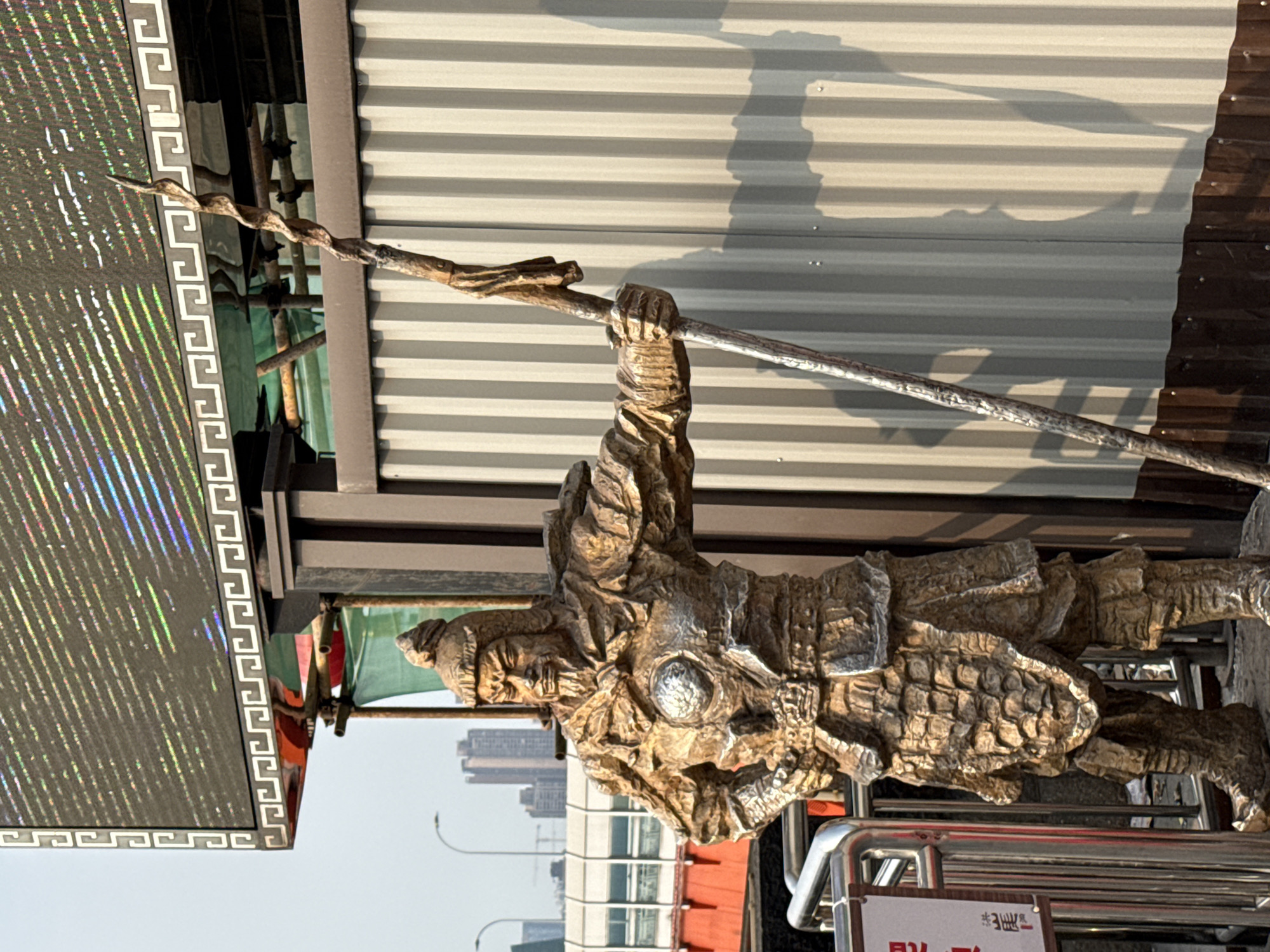 A bronze sculpture of a warrior in armor holding a pole is installed along the side of a building on a street in Yuzhong District, China. The pole he is holding is resting on a wooden support beam. In the background is a building complex.