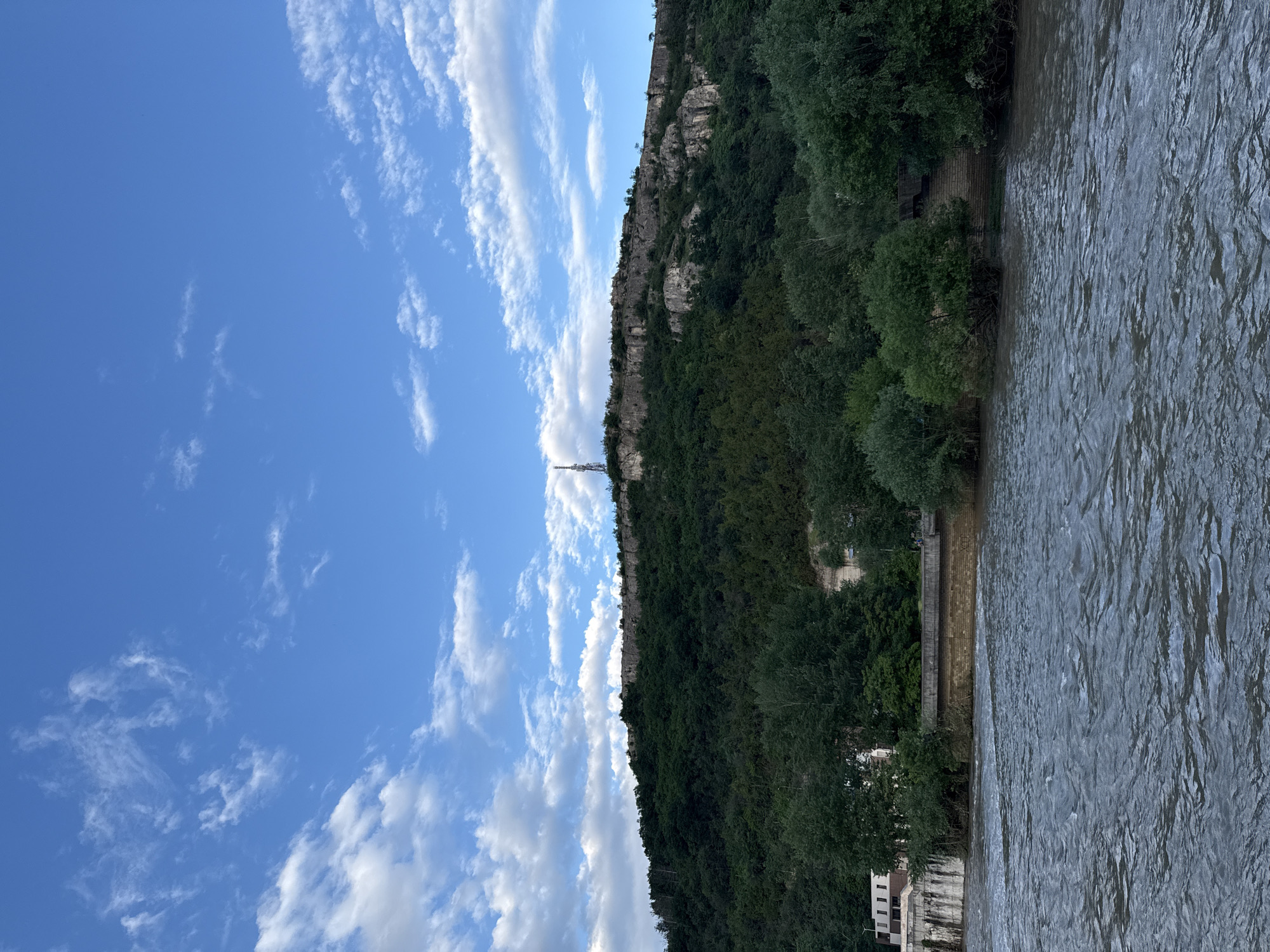 The image shows a section of the Osam River in Lovech, Bulgaria. The river flows on the right side of the frame with visible ripples on its surface. On the left side of the river is a bank lined with trees and some buildings visible at the bottom. A large, steep hill covered in dense vegetation rises prominently in the center of the image. A telecommunications tower stands on the hill. The sky occupies the upper left portion of the image, displaying a bright blue color with scattered white clouds. The orientation of the photo is rotated 90 degrees to the right.