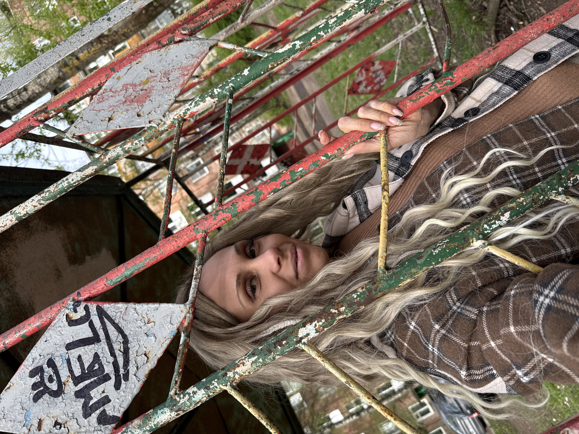 A woman with long blonde hair is positioned behind a decaying, multi-colored metal fence. She is looking directly at the camera with a slight smile. She is wearing a brown and white checkered jacket over a brown top. The fence, which has peeling paint in red, green, and white, obstructs a partial view of a building in the background. A diamond-shaped sign with the partially obscured word "SHELL" in black lettering is visible on the fence. The photo was taken in the city of Rusanovo, Russia.