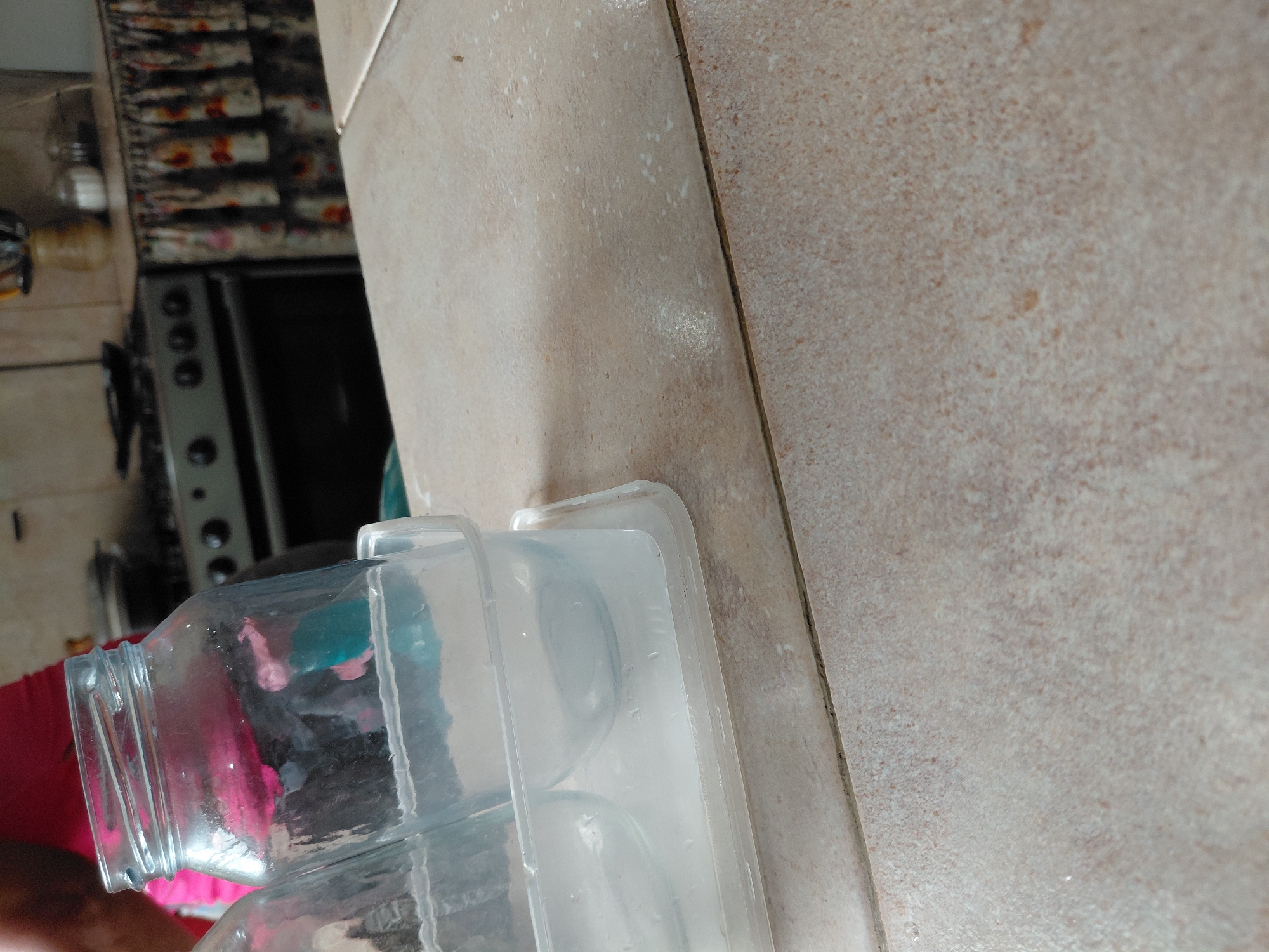 This close-up, tilted image captures a section of a kitchen counter in what appears to be a home in Santa Ana, Venezuela. The main focus is on two clear containers resting on a light-colored tiled surface.
On the left, a transparent glass jar, possibly for storage, is lying horizontally. Beside it, a clear plastic container, resembling an ice cube tray with circular depressions, is also on its side, appearing to contain water or ice.
A person wearing a red garment is partially visible on the far left, their arm or shoulder area cropped into the frame.
The background, significantly blurred, reveals more of the kitchen environment. A black and silver stove or oven unit with visible control knobs is positioned in the lower-left background. Above the stove, there's a patterned textile or wall covering featuring dark and rust-colored designs. Several small jars or containers, possibly for spices or condiments, are discernible on a shelf or counter further back.
The countertops are made of light beige or cream-colored ceramic tiles, showing visible grout lines and some speckling. The overall lighting suggests it is daytime. There is no specific activity overtly taking place; the items are stationary on the counter. No text is visible in the image.