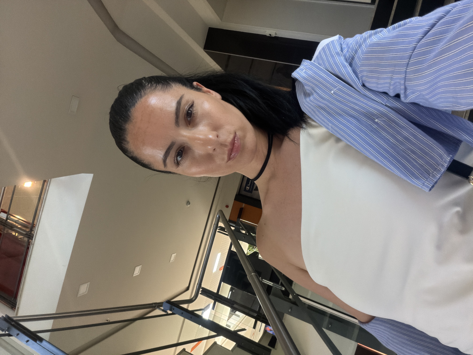 A medium shot shows a woman posing for a selfie indoors, likely in the lobby or a common area of a building with a modern architectural design. She is standing on a staircase, with a sleek metal railing and glass partitions visible behind her. The woman has her dark hair pulled back in a ponytail and is wearing a white, off-the-shoulder top and a blue and white striped shirt draped over her shoulders. She is wearing a black choker necklace. The background reveals a multi-story interior with neutral-colored walls and ceiling, and recessed lighting. The overall impression is of a contemporary space, possibly a hotel or commercial building, in the city of Pleven, Bulgaria.