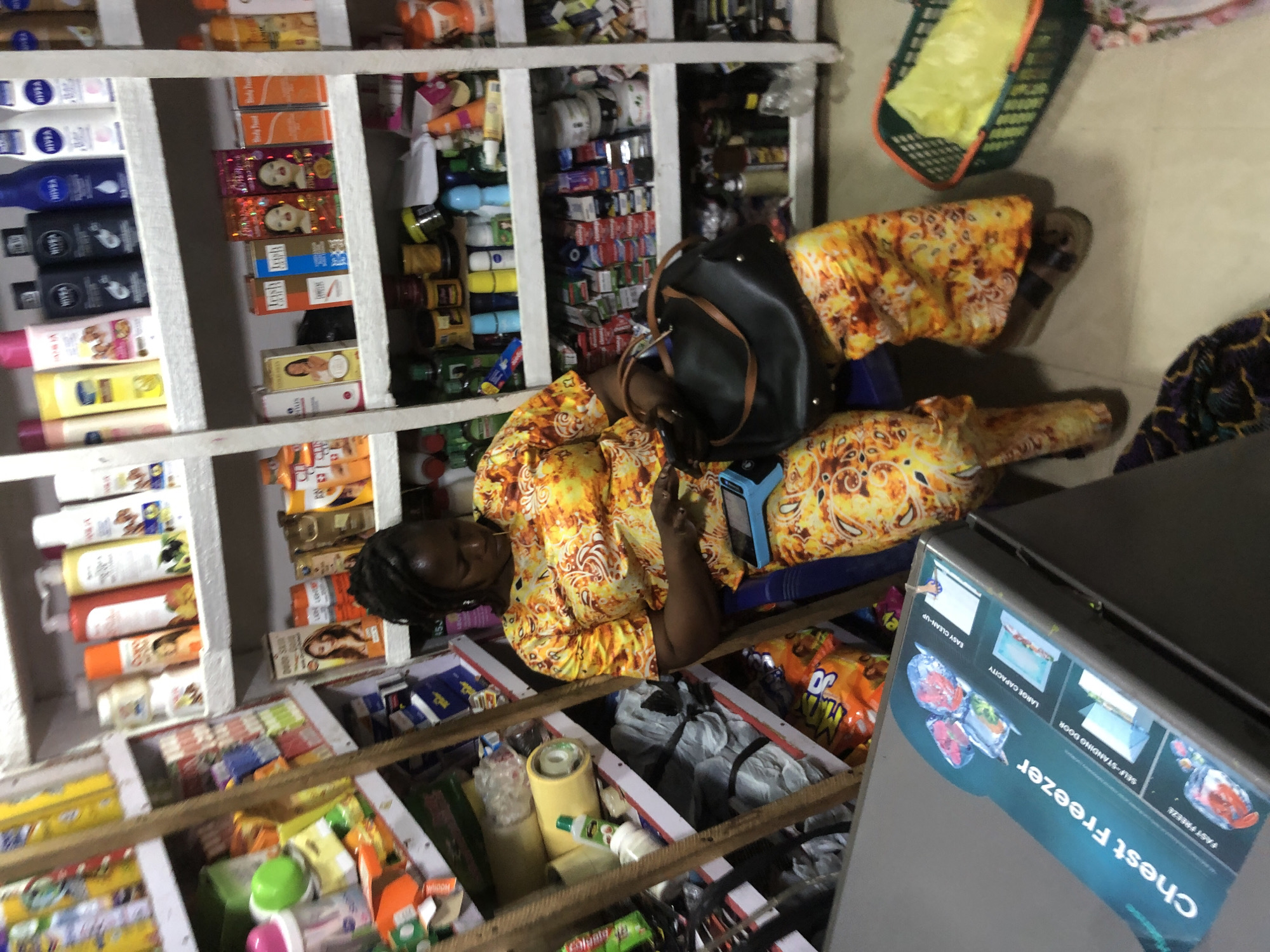 The image shows a woman sitting in a store filled with shelves of products, likely in Ado, Nigeria. The woman is wearing a yellow and orange patterned outfit and is looking down at a device in her hands. A black handbag is resting on her lap. The shelves are stocked with various items, including toiletries, food products, and cleaning supplies. There is also a chest freezer visible in the foreground with a label indicating "Chest Freezer." The overall scene depicts a retail environment with a customer engaged with her phone.
