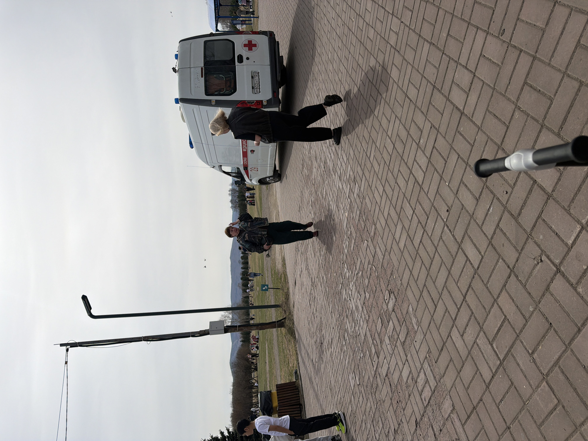 The image depicts an outdoor scene on a brick-paved pathway in Krasnoyarsk, Russia. An ambulance is parked near the pathway. A woman with blonde hair is walking away from the ambulance. Another person is walking on the pathway towards a park. In the park there are trees, a trash can, people and a road. A boy is performing a push-up in the foreground. A piece of equipment (possibly a camera stand) is also in the foreground. In the background there are mountains. The sky is overcast.