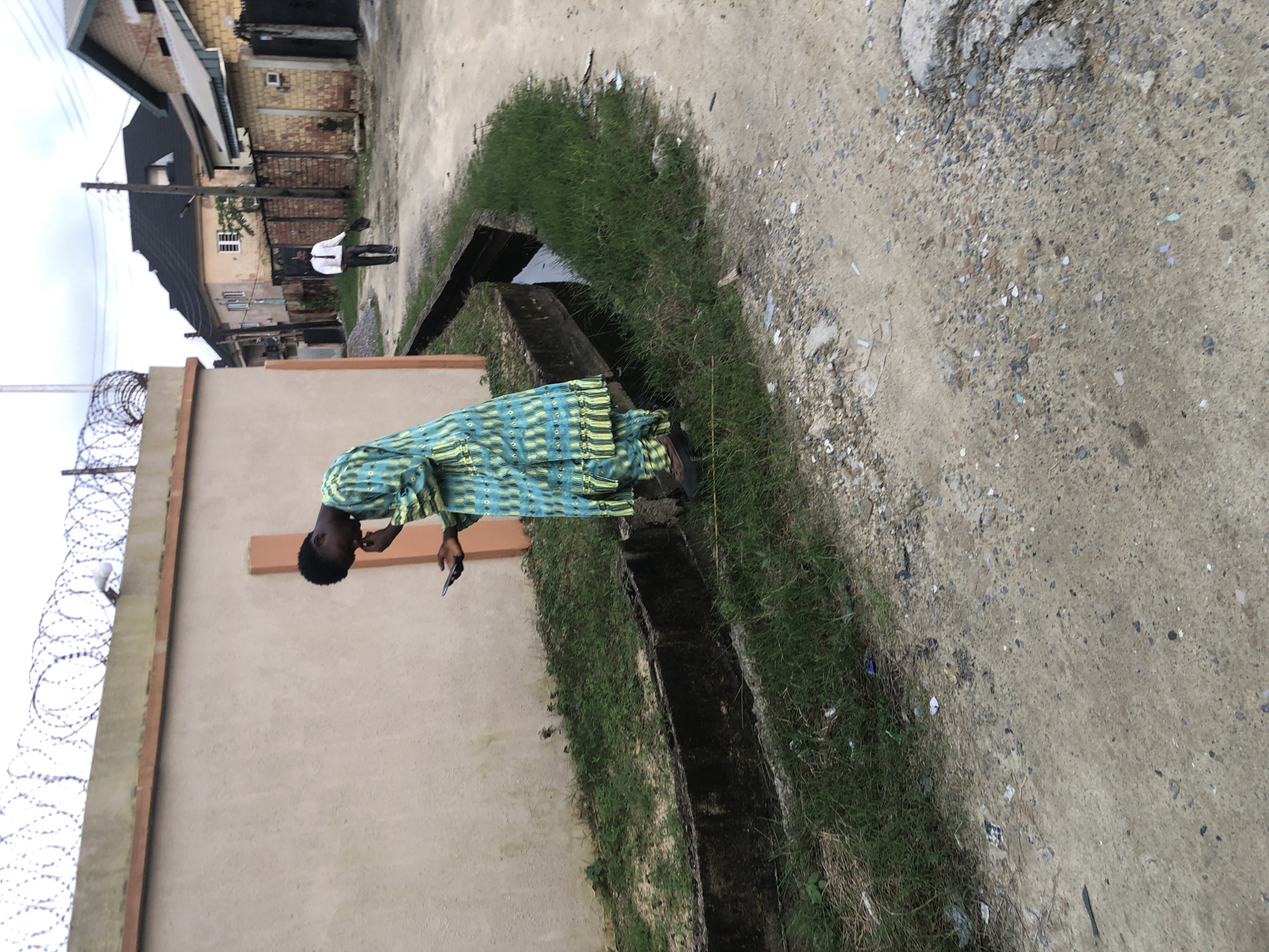 In Ado, Nigeria, a man wearing a blue and green patterned traditional outfit stands next to a drainage ditch, looking down at his phone. His right hand is raised towards his face, obscuring part of it, while his left hand holds the phone. Behind him, a rough dirt road leads past houses with corrugated metal roofs and fenced compounds. Further down the road, another man in a white shirt and dark trousers walks away, carrying a dark bag. The scene appears to be in a residential area, with a concrete wall topped with barbed wire running along the left side of the image. The ground is uneven, with patches of grass and scattered debris. The sky is overcast.