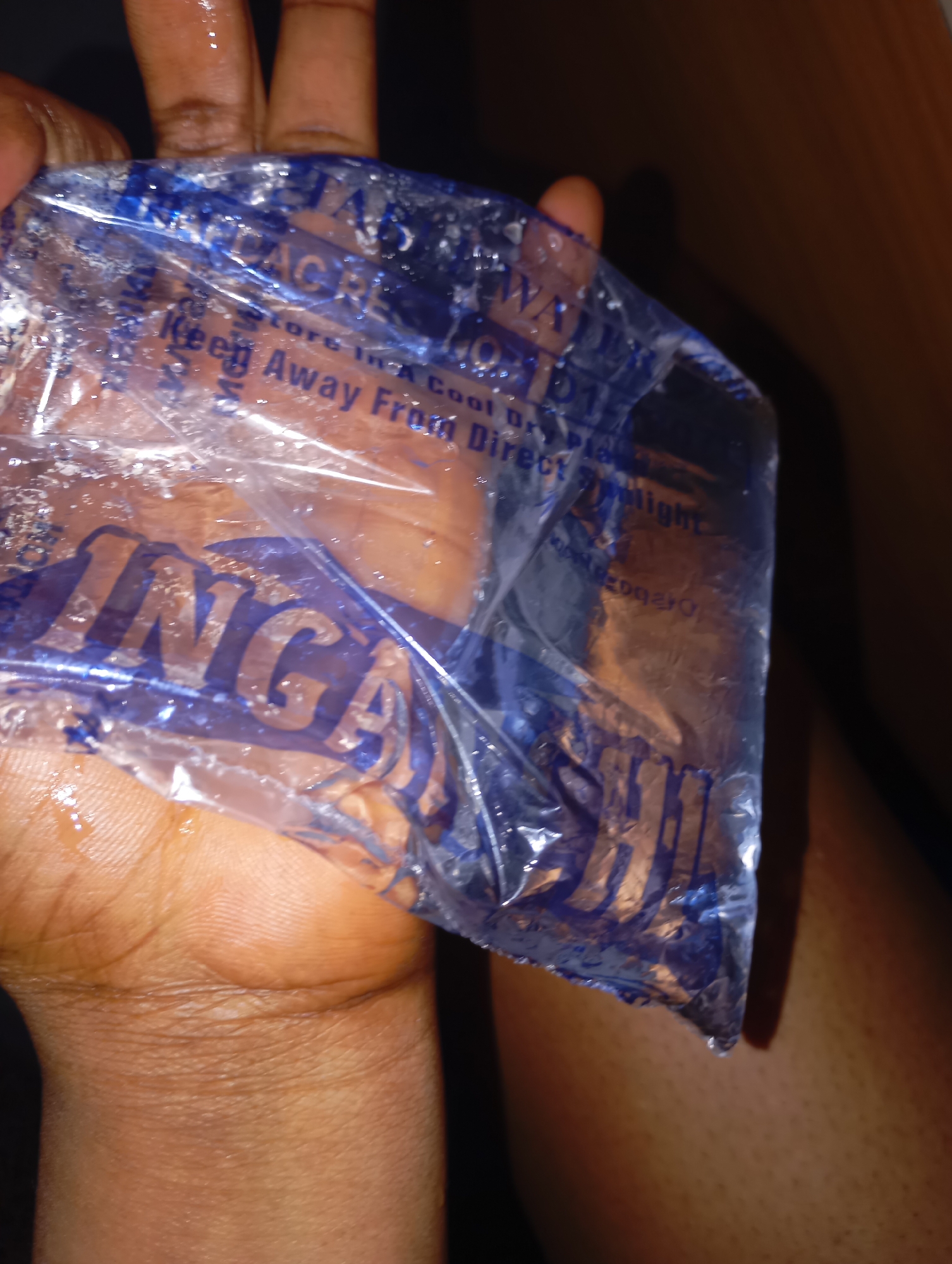 A hand is holding a clear plastic bag filled with water. The bag has blue text on it, some of which is legible. One prominent word appears to be "INGER". There are water droplets on the outside of the bag and on the hand. The background is dark and out of focus, with a suggestion of skin tone in the upper right and lower right of the image.