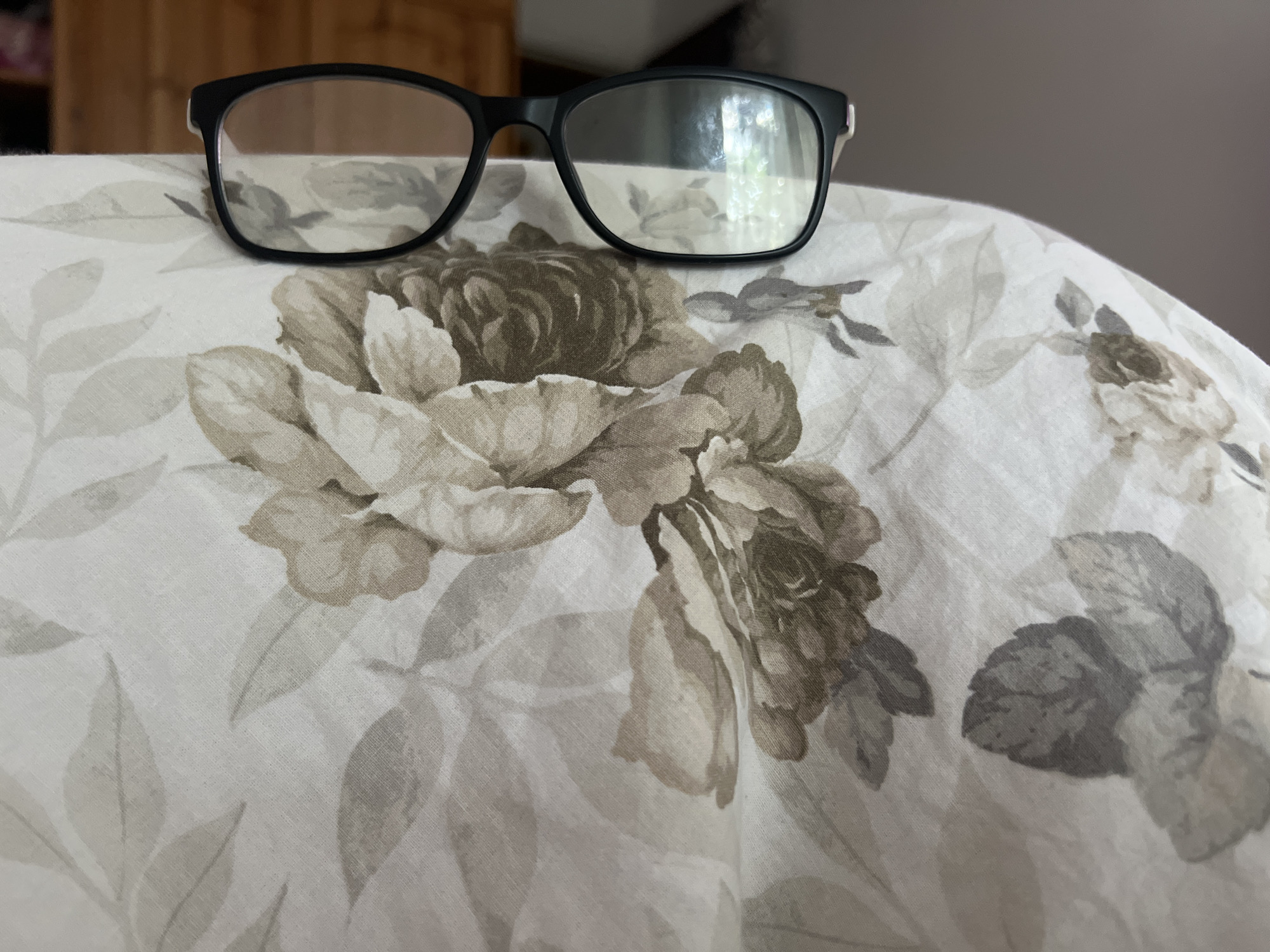 The image shows a pair of black-rimmed eyeglasses resting on a fabric surface with a floral pattern. The fabric appears to be a light color with muted brown and gray floral designs. The eyeglasses have clear lenses and are positioned centrally in the upper portion of the frame. The background is softly blurred, suggesting an indoor setting, possibly a bedroom or living room, with wooden furniture visible in the upper left corner. There is no indication of the city of Nalchik, Russia, or any specific location within it. The scene is static and depicts everyday objects.