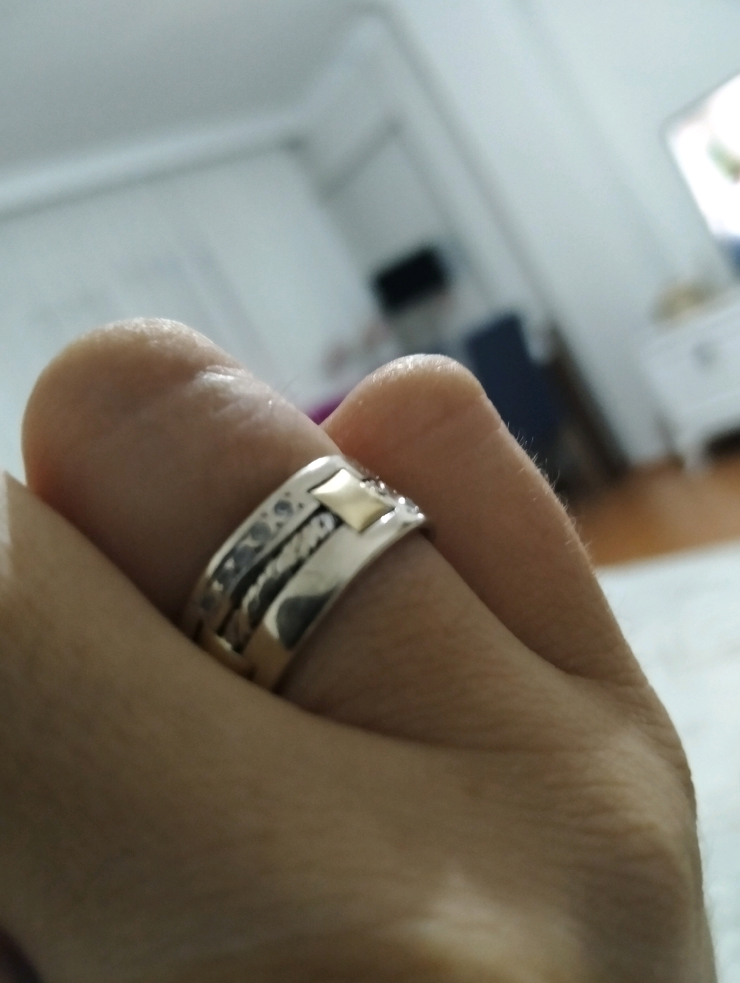 A close-up, angled shot captures a hand with a silver ring featuring textured bands and a gold square accent. The ring is worn on the index finger, which is partially visible against a blurred background of what appears to be an indoor setting, possibly in Balçova, Turkey. The background includes indistinct furniture and lighting, suggesting a domestic or commercial space. The lighting is soft, with some highlights on the ring and skin. The focus is primarily on the ring and the finger.