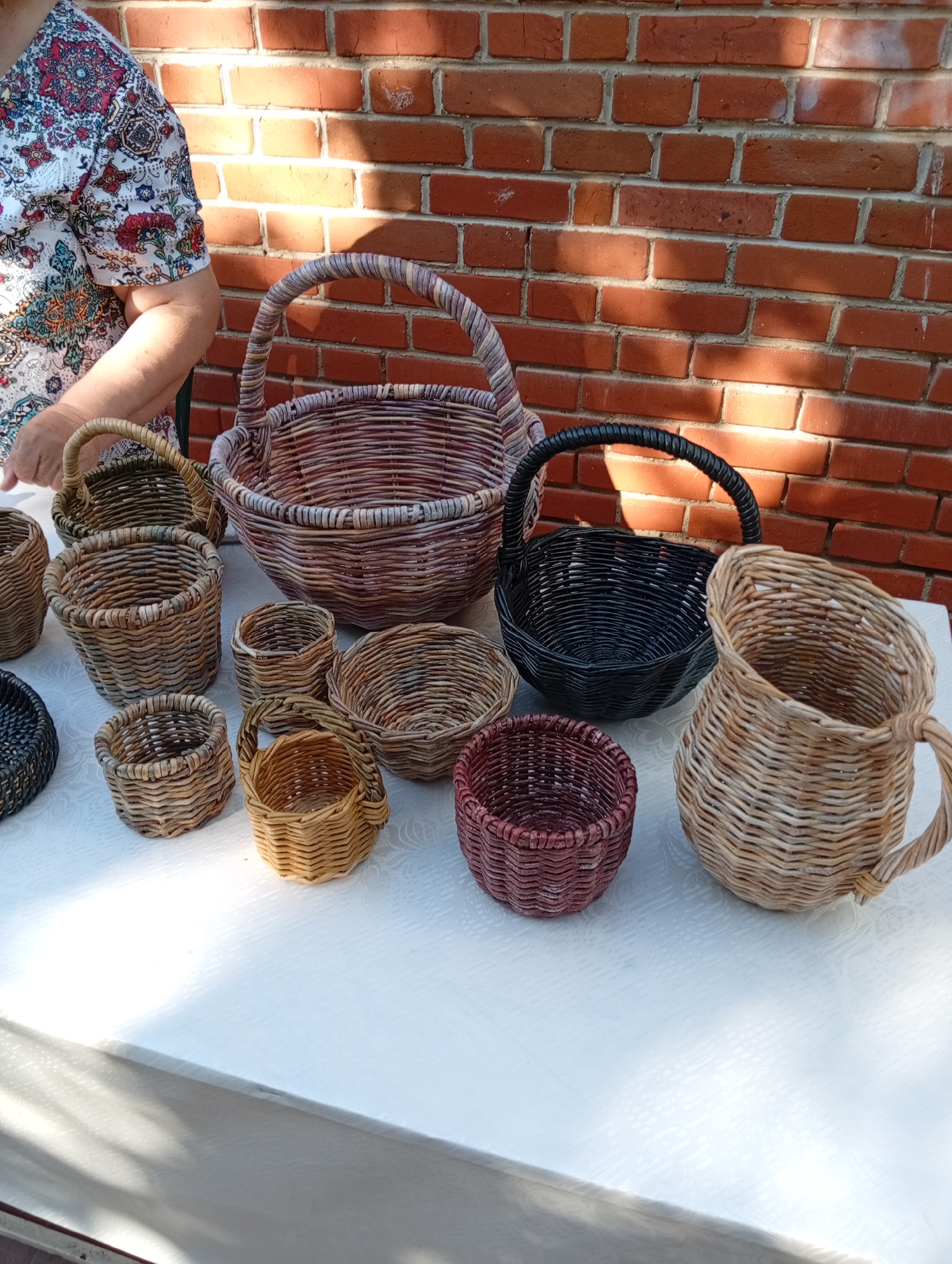 A still life photograph captures a collection of woven wicker baskets displayed on a white tablecloth in front of a red brick wall. The baskets vary in size and color, with some in natural wicker tones, others in dark brown, and one a muted purple. Several of the baskets have handles. In the background, a portion of a person wearing a patterned shirt is visible, their arm resting on the table. The scene is lit by natural sunlight, casting shadows on the brick wall and the table. This display appears to be set up outdoors, possibly at a market or craft fair, with the brick wall suggesting a building in the background. The overall impression is one of handmade crafts being showcased. The photographer's metadata does not provide location information; however, the provided context places this scene in the city of Kostanay, Kazakhstan.