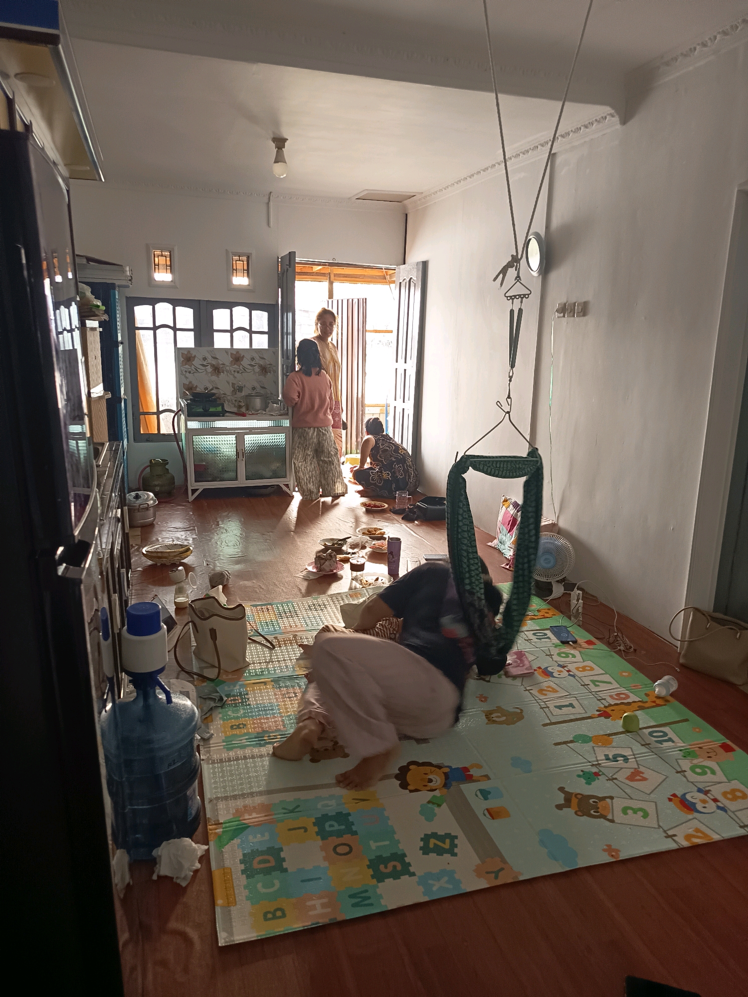 The image depicts an indoor scene, likely a residential dwelling in Banjarmasin, Indonesia. The room appears to be a living area and kitchen combined. The floor is wooden, with a colorful educational mat laid out. In the foreground, a person is lying down on the mat, with their legs extended. Nearby is a large blue water dispenser. To the left, part of a black refrigerator is visible.
In the mid-ground, two women are standing near a kitchen counter which is equipped with a stove and some utensils. Another person is seated on the floor to the right, facing away from the camera, with plates of food arranged around them. A green fabric swing is suspended from the ceiling by ropes. A small electric fan is also visible.
The overall lighting suggests it is daytime, with natural light entering from an open doorway or window in the background. The walls are painted white, and decorative molding is present along the ceiling. The environment appears to be a modest, lived-in space.