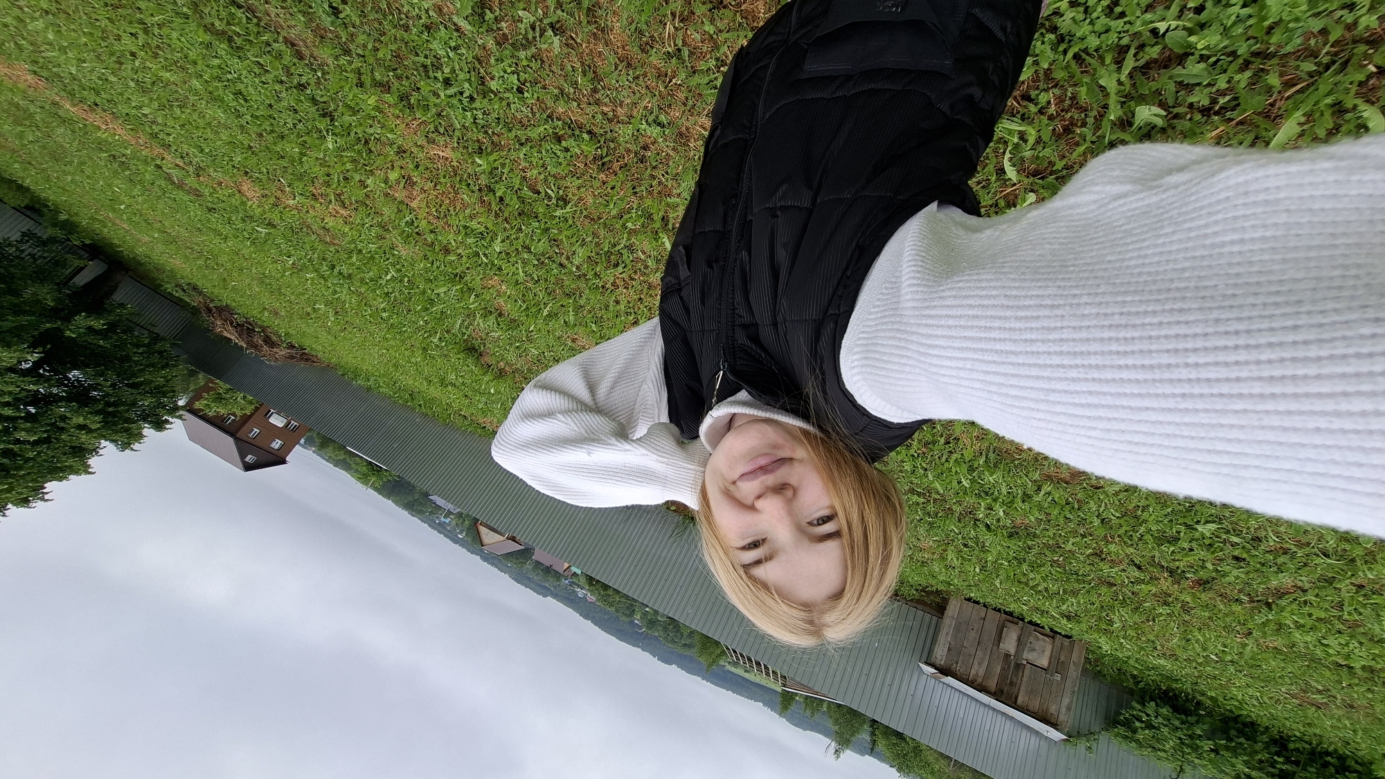 A fair-skinned woman with blonde hair takes a selfie in Большой Букор, Russia. She wears a white sweater and a black vest. The background includes a green lawn, a fence, houses, and a cloudy sky.