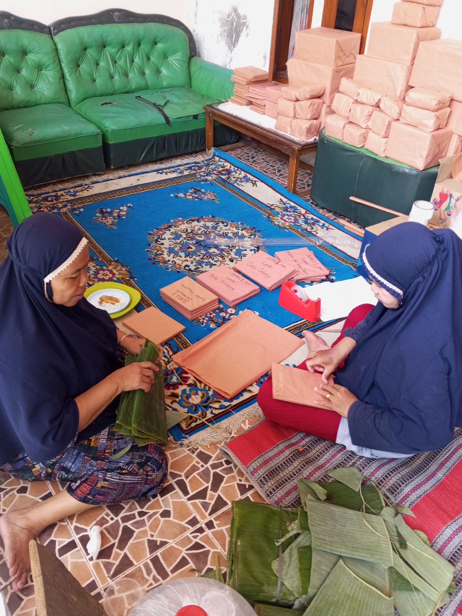 In a domestic setting in Ciruas, Indonesia, two women are seated on a patterned rug, engaged in food preparation. The woman on the left, wearing a dark blue hijab and patterned clothing, is meticulously wrapping what appears to be food items in banana leaves. In front of her, a small plate holds a few cooked food items. The woman on the right, also in a dark blue hijab and red trousers, is folding a light brown paper package, suggesting she might be preparing to wrap the banana leaf-wrapped items or other goods.
The environment is furnished with a green tufted sofa and a wooden bench piled high with numerous light brown paper-wrapped packages, likely gifts or goods for sale. A blue rug with an intricate floral pattern covers the floor. Scattered around are additional pieces of brown paper and a roll of tape. The room appears to be a residential interior, possibly a living area, where these activities are taking place. The overall scene depicts a communal effort, perhaps for an event or business.