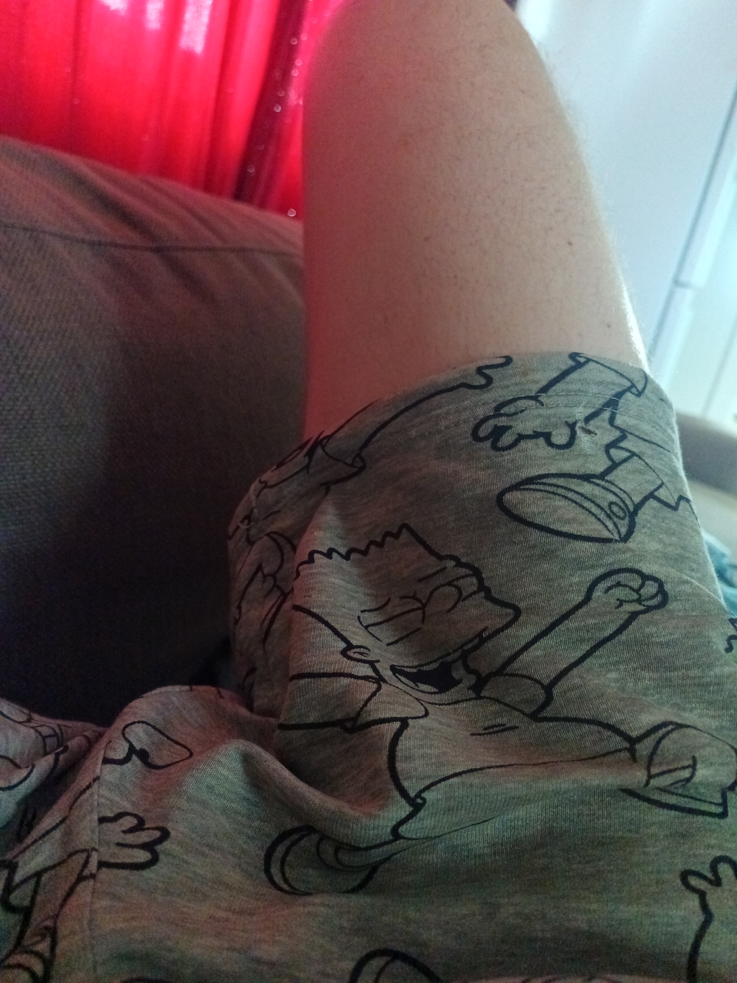 The image is a close-up shot of a person's leg wearing grey pajama bottoms with a cartoon print. The print appears to be of Bart Simpson and other characters from The Simpsons. The leg is resting on a dark-colored surface, possibly a couch or chair, and a red, translucent fabric, likely a curtain, is visible in the background. The lighting suggests an indoor setting. The image does not contain any information about the location being Risøyhamn, Norway.