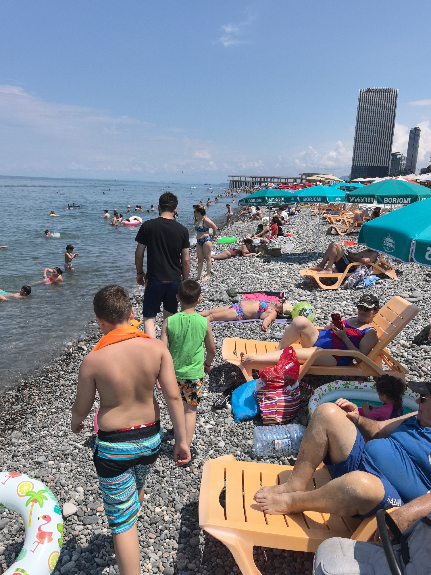 This is a wide-angle, eye-level shot of a crowded beach on a sunny day. The foreground is dominated by a pebbly beach with several people relaxing on orange plastic lounge chairs. In the middle ground, the sea is filled with swimmers and inflatable toys. The background shows a pier with more people, and beyond that, tall buildings and a few boats on the horizon. The sky is clear blue with a few scattered clouds. Several bright turquoise beach umbrellas are visible, some bearing the "BORJOMI" logo.
In the immediate foreground, the backs of two young boys are visible, one wearing an orange towel around his neck and patterned swim shorts, the other in a green t-shirt and patterned swim shorts. To the left, a boy's legs and patterned swim shorts are visible, along with a flamingo-themed inflatable ring.
Further back, several people are sunbathing or sitting on lounge chairs. One woman in a blue and white bikini is sitting up and looking at her phone, while another woman in a patterned bikini is lying down. A man in a black t-shirt and blue shorts walks away from the camera towards the water, and a woman in a bikini is standing near him.
In the water, various people are swimming, including children and adults. Some are using inflatable rings. The water is calm and clear. The beach is covered in smooth, grey pebbles. The overall atmosphere is one of a busy summer day at the beach.