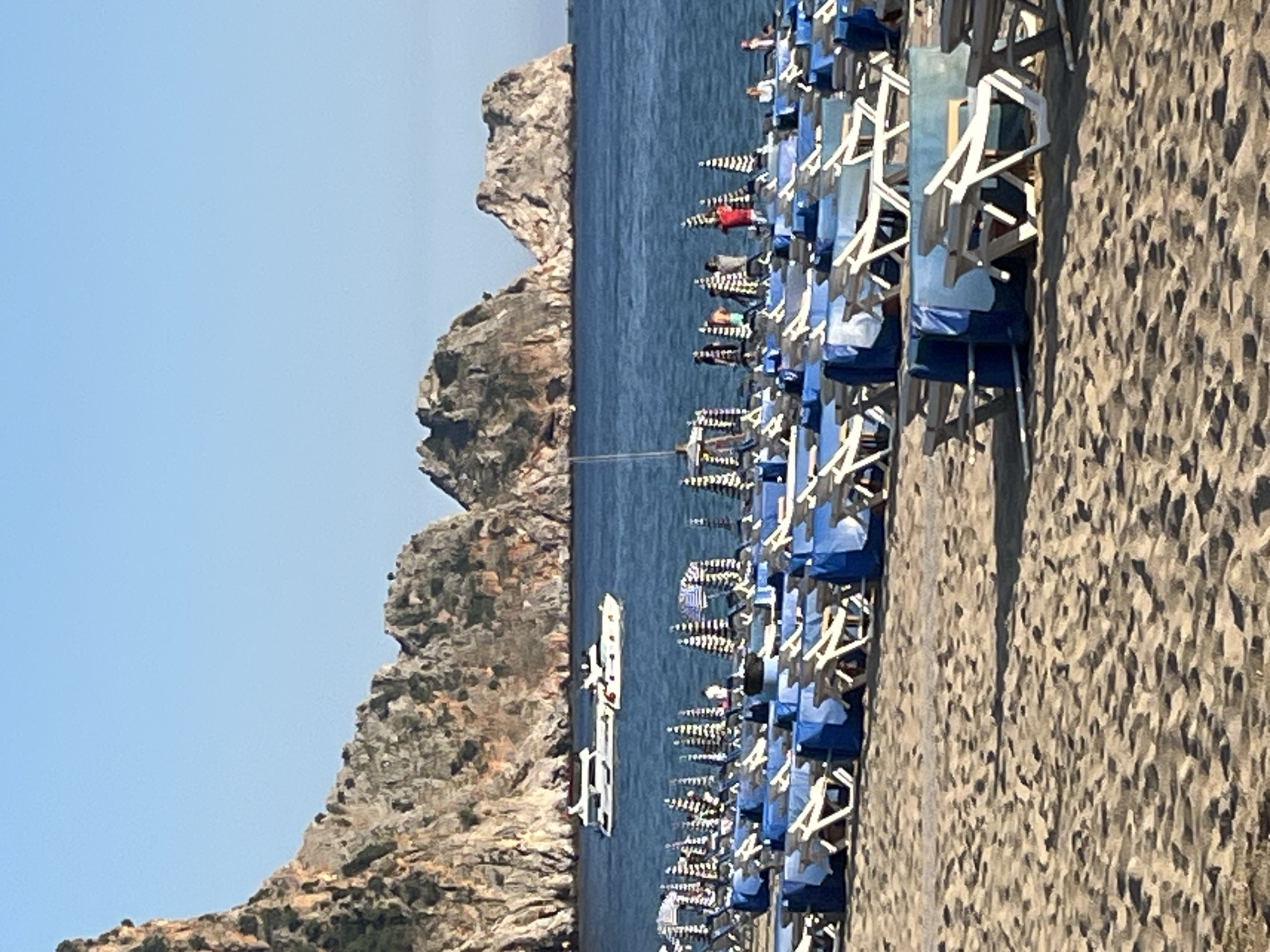 The image depicts a coastal scene at Kleopatra Beach in Alanya, Turkey. A rocky promontory is visible to the left, with the blue sea stretching between it and the beach. A boat is in the water. The beach is lined with rows of closely packed beach chairs, mostly blue and white, many of which are occupied by people. Fine-grained sand covers the beach. The sky is clear and blue.