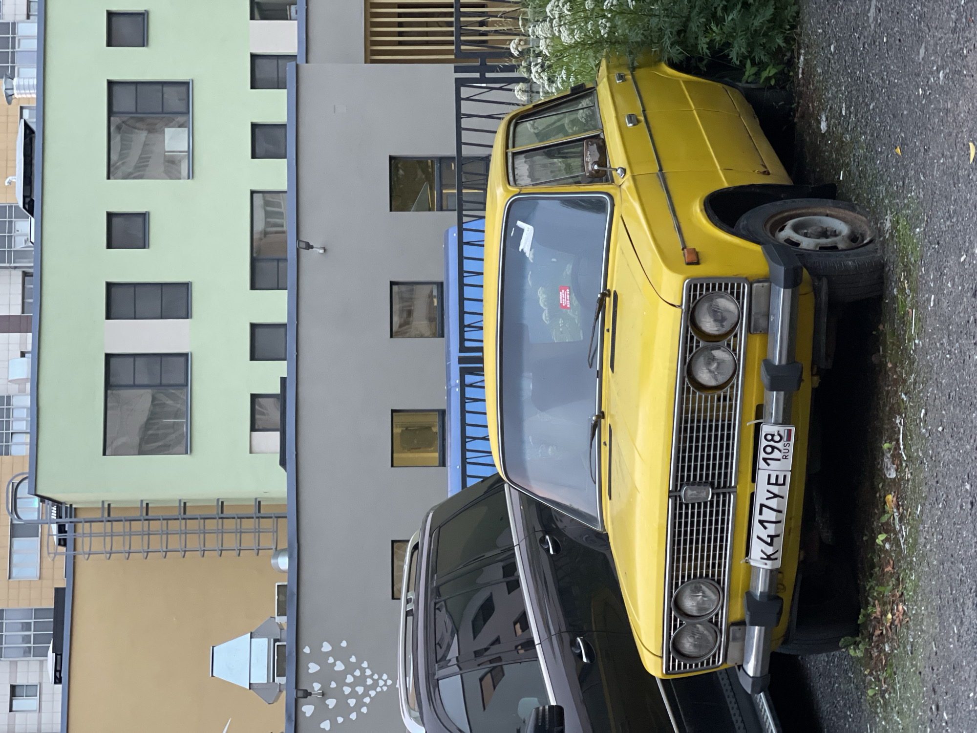 A bright yellow vintage car, possibly a VAZ-2101 "Zhiguli," is parked on a gravel surface in front of a modern building. The car has a chrome grille, round headlights, and a license plate that reads "K417YE 198 RUS." To the left of the yellow car is a dark-colored SUV. The building in the background is multi-colored, featuring sections of pale yellow, light green, and grey. The light green section of the building has several dark-framed windows. A metal fire escape is visible on the pale yellow section. White heart-shaped drawings are present on the grey portion of the building above the cars. White flowers are growing to the right of the yellow car, partially obscuring its rear.